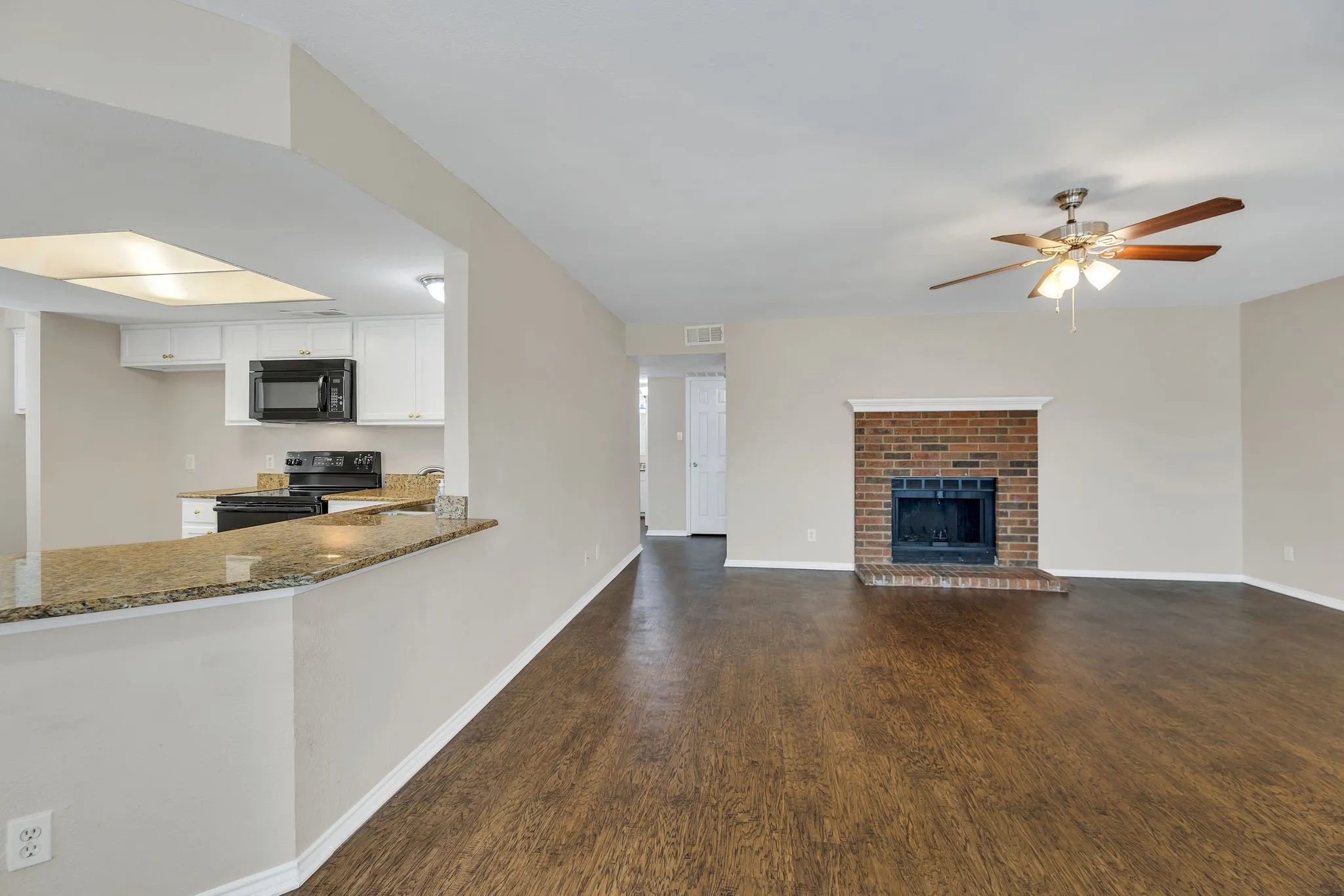 Unit C- Wood-finish flooring throughout, brick fireplace with mantel, white kitchen cabinetry, granite-style countertops, and a ceiling fan with integrated lighting