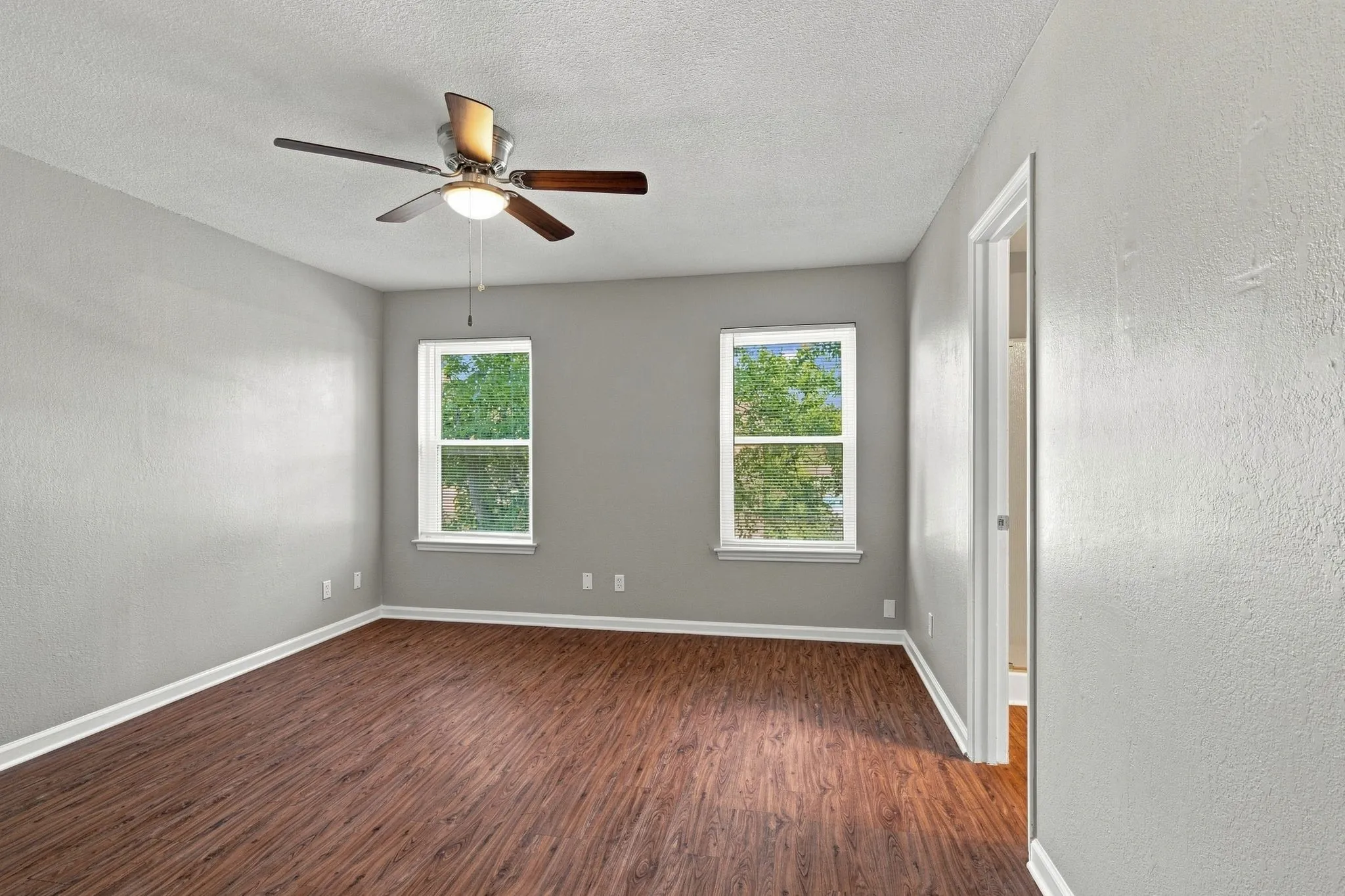 Unit D -Wood-finish flooring throughout, two windows, ceiling fan with integrated lighting, white trim, and a neutral wall color