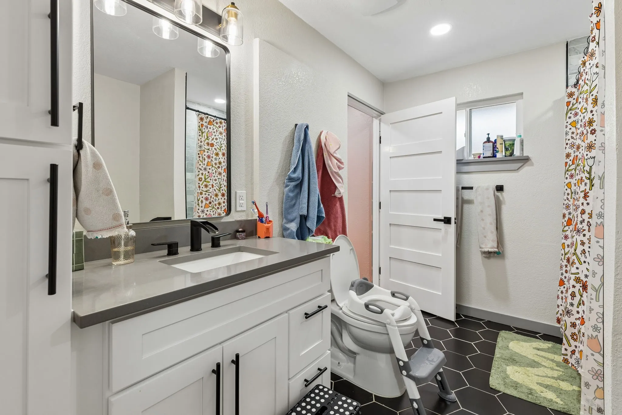 White vanity with a light gray countertop and black fixtures, framed mirror with overhead vanity lighting, dark hexagonal floor tiling, and a white interior door with black hardware
