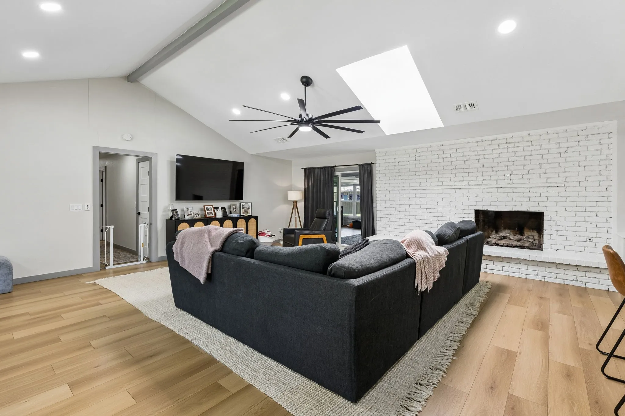 Vaulted ceiling with exposed beam, wood-finish flooring, white-painted brick fireplace, recessed lighting, and a skylight