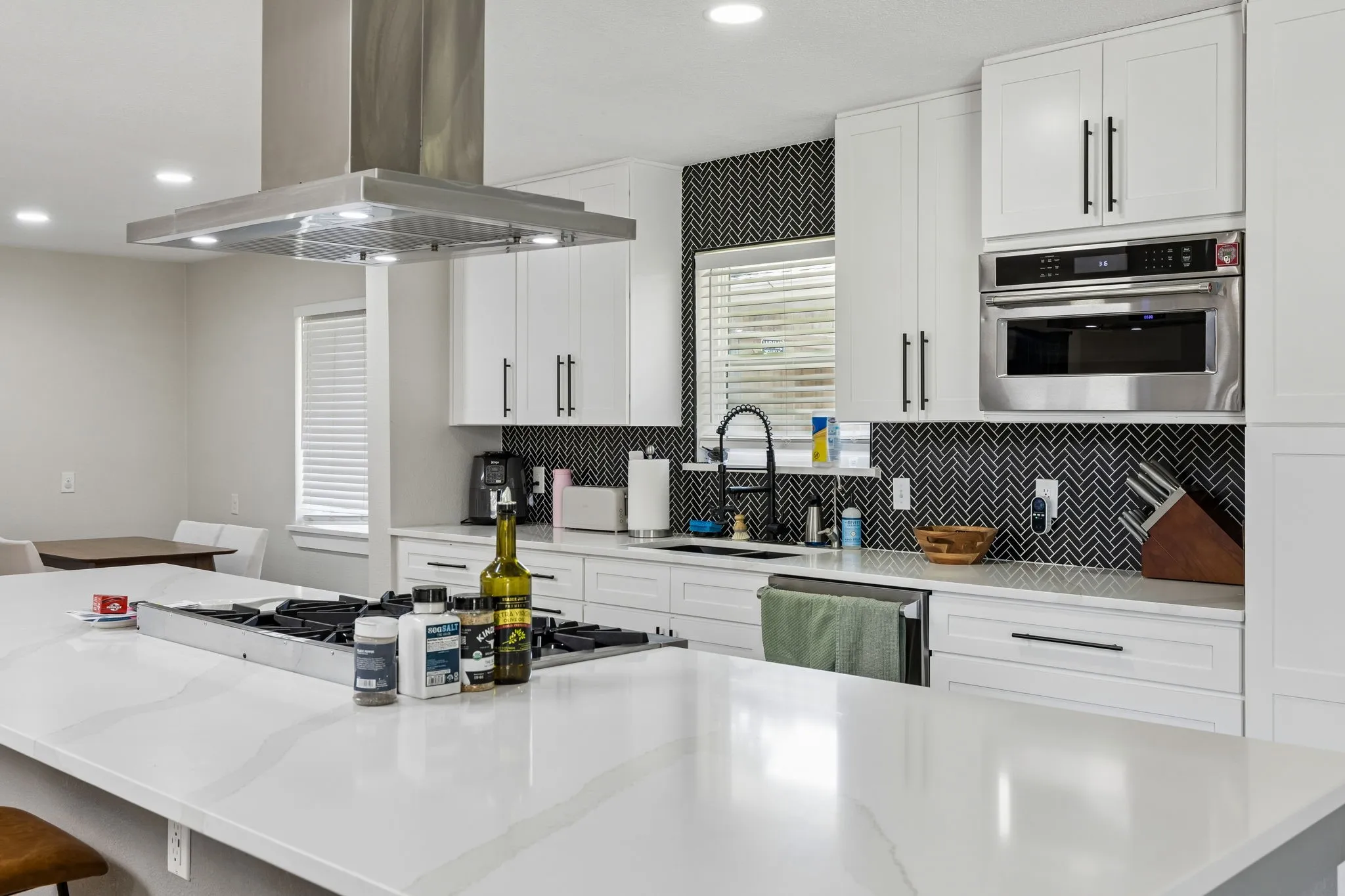 Kitchen featuring a central island with a built-in cooktop, stainless steel range hood, white shaker cabinetry with dark hardware, and a black herringbone tile backsplash