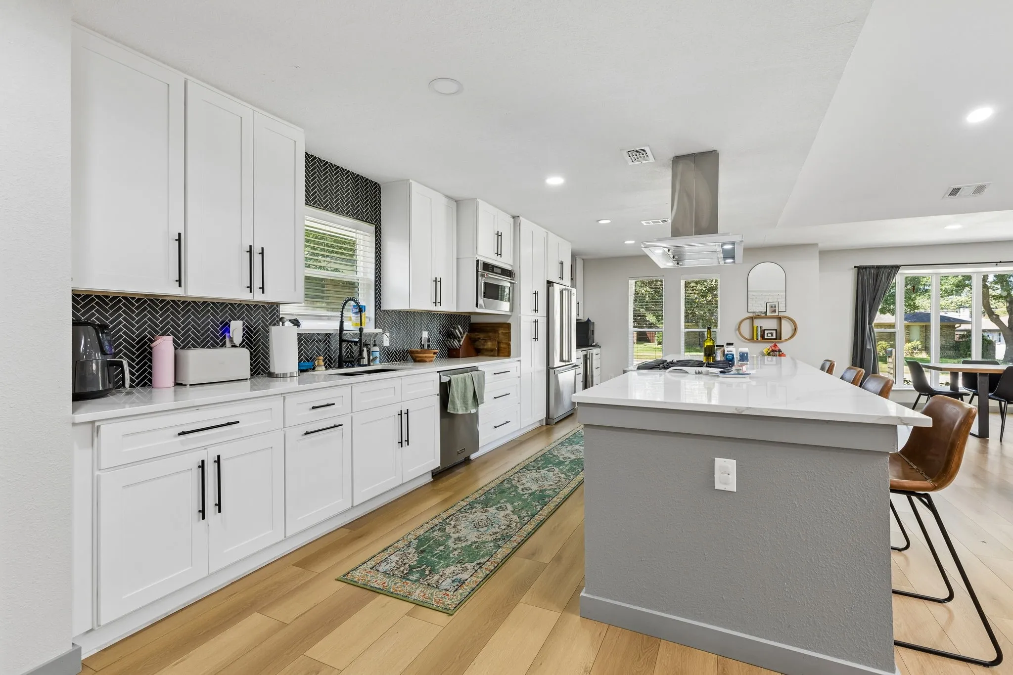 Contemporary kitchen featuring white cabinetry, a black herringbone tile backsplash, an expansive island with a light-toned countertop, and wood-finish flooring