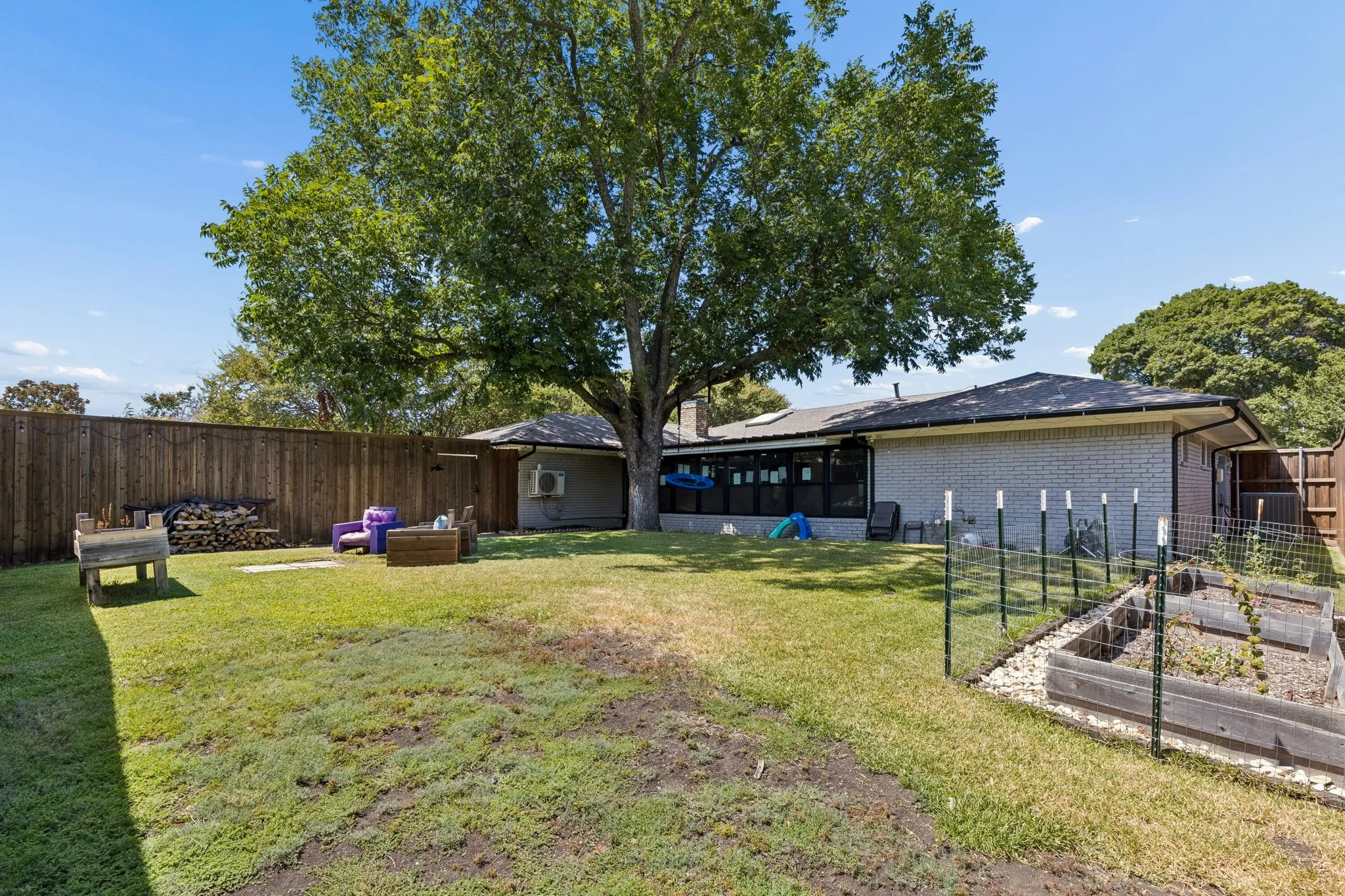 Expansive backyard featuring a mature tree, wood privacy fencing, light-colored brick exterior, and raised garden beds