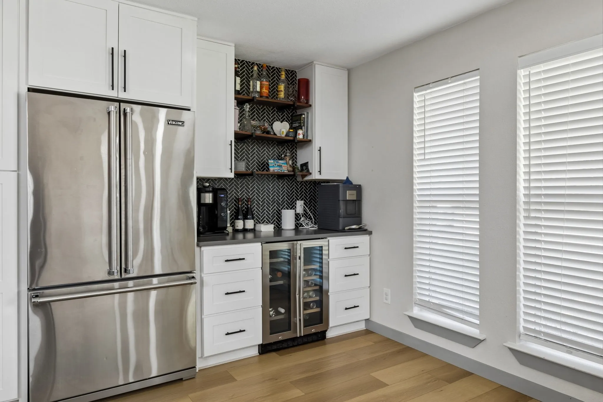 Stainless steel refrigerator, white shaker-style cabinetry, built-in beverage cooler, dark countertops, wood-finish flooring