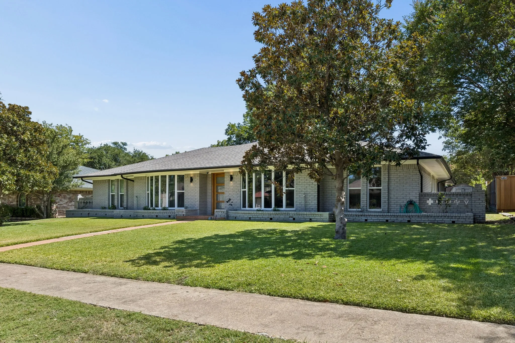 Gray brick exterior with a dark shingle roof, a front walkway, and mature trees