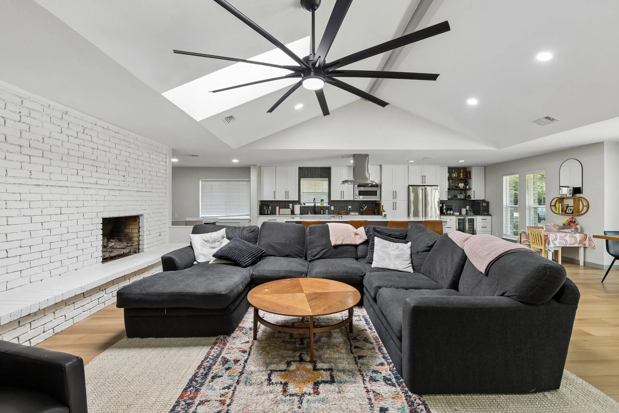 Vaulted ceiling with skylight and large ceiling fan, white painted brick fireplace, wood-finish flooring, and recessed lighting