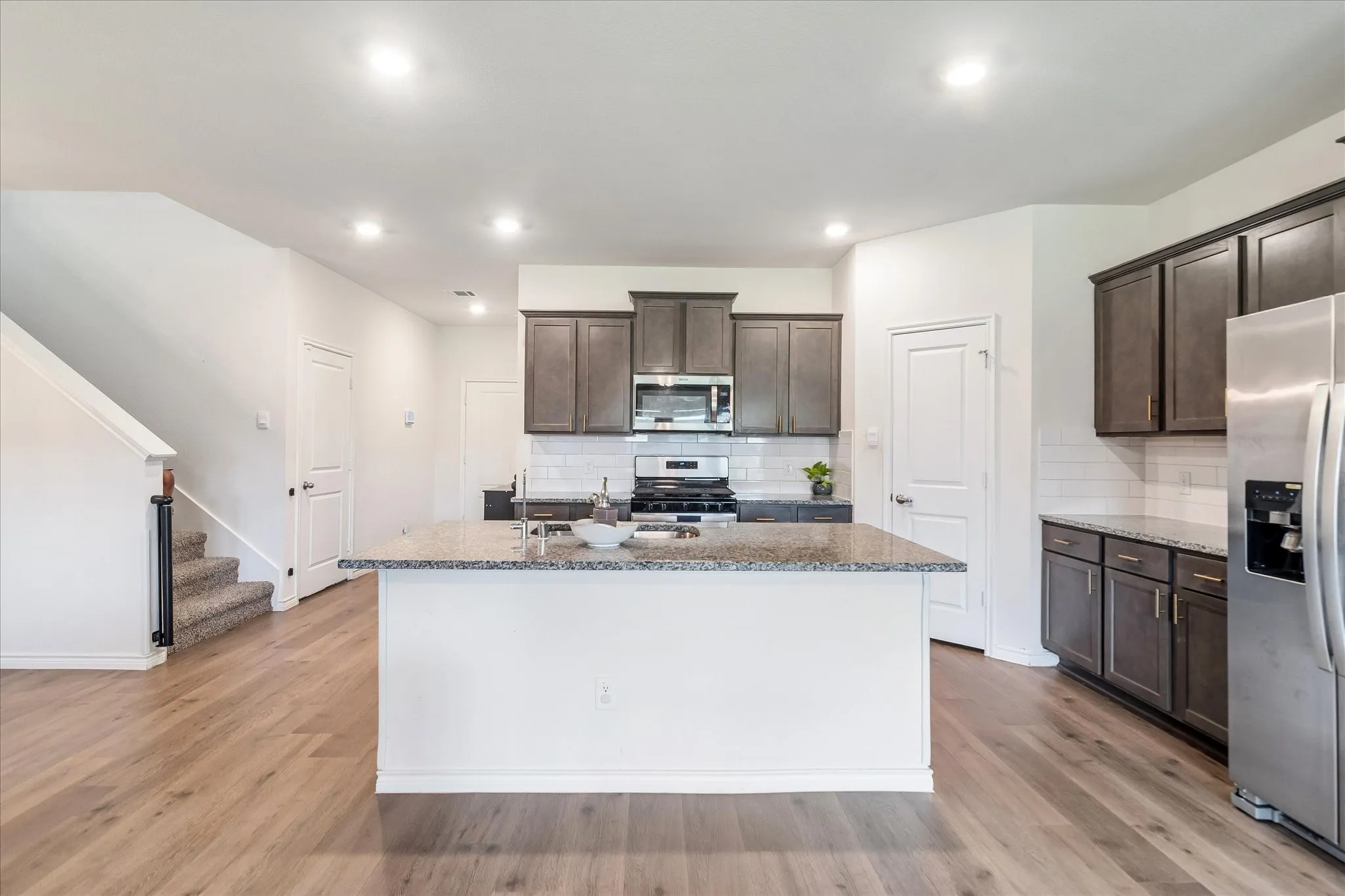 Kitchen featuring an island with a sink, stainless steel appliances, dark wood-finish cabinetry, subway tile backsplash, and light wood-finish flooring