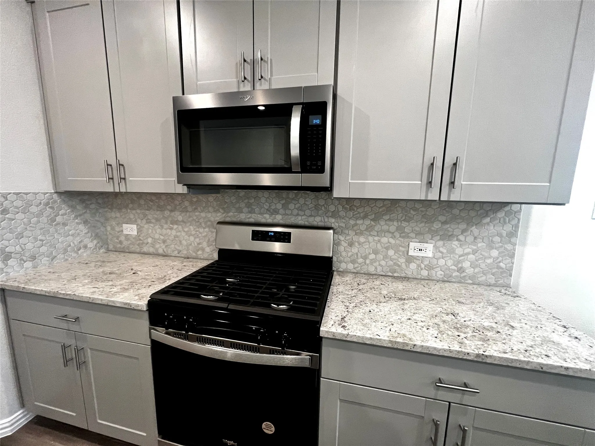 Light gray cabinetry with brushed nickel hardware, stainless steel microwave, stone mosaic backsplash, and granite-patterned countertops