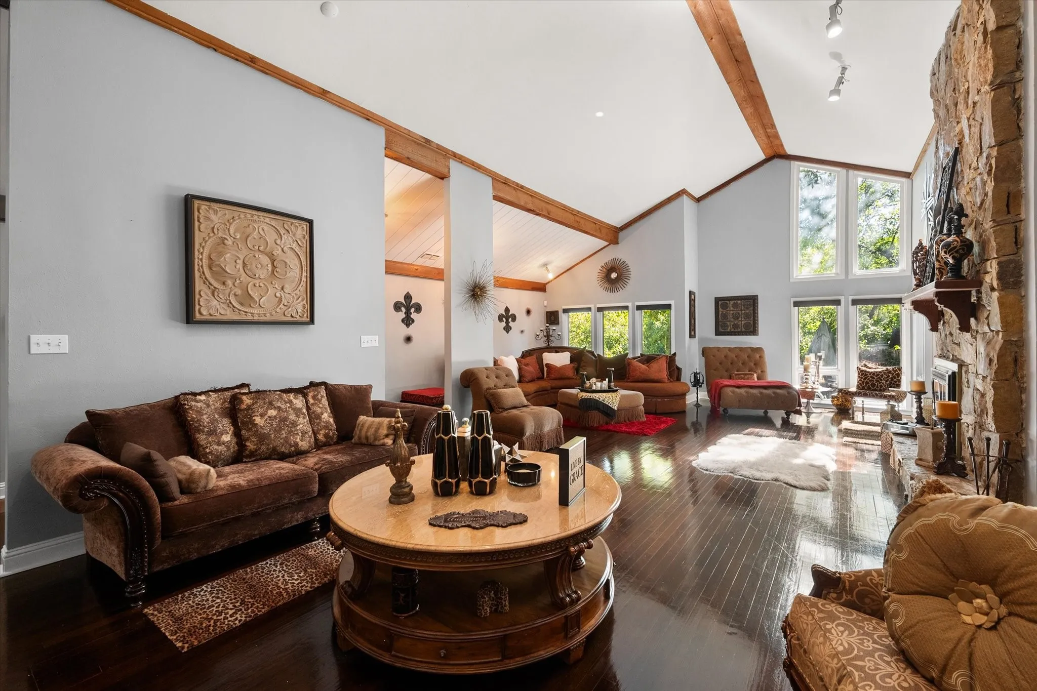 Vaulted ceilings with exposed wood beams, expansive windows, dark wood-finish flooring, and a stone-clad fireplace