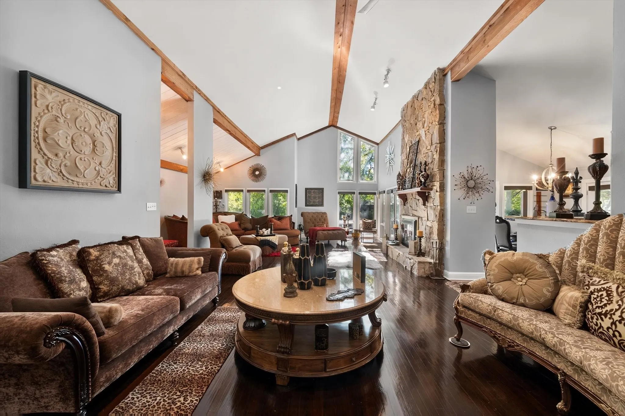 Vaulted ceilings with exposed wood beams, a stone-clad fireplace, dark wood-finish flooring, and large picture windows