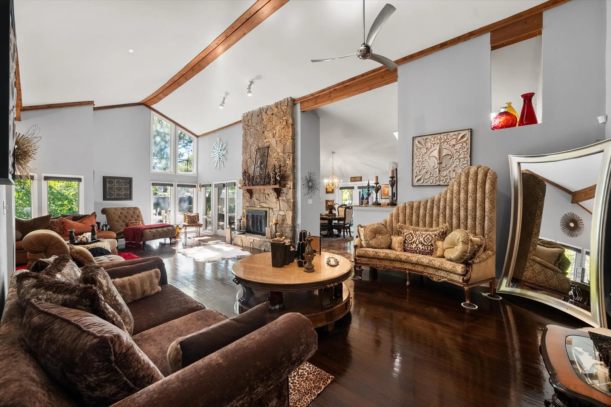 Vaulted ceilings with exposed wood beams, a stone-clad fireplace, large windows, and dark wood-finish flooring