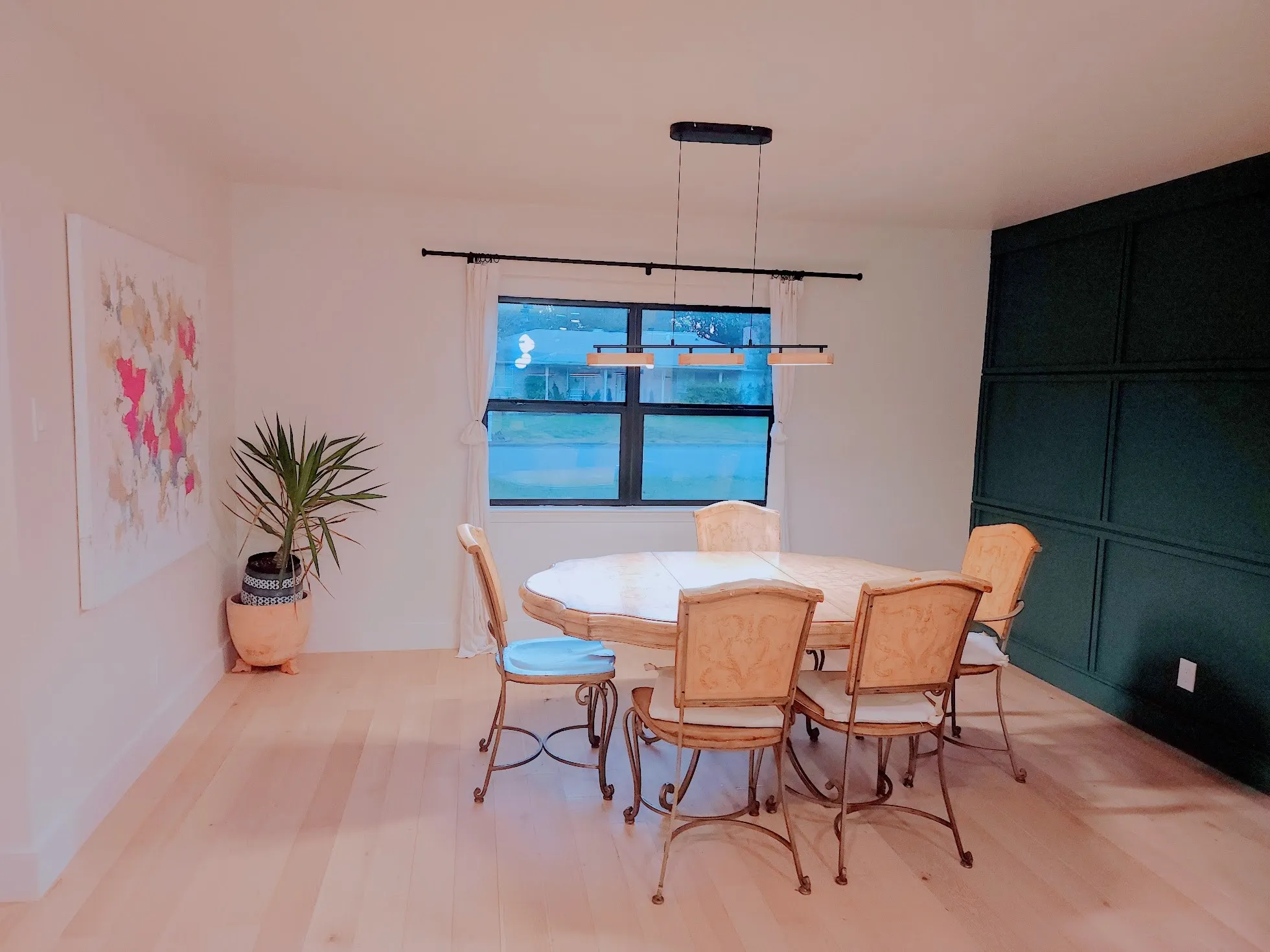 Light wood-finish flooring, accent wall with wainscoting, window with white drapery, and a linear pendant light fixture