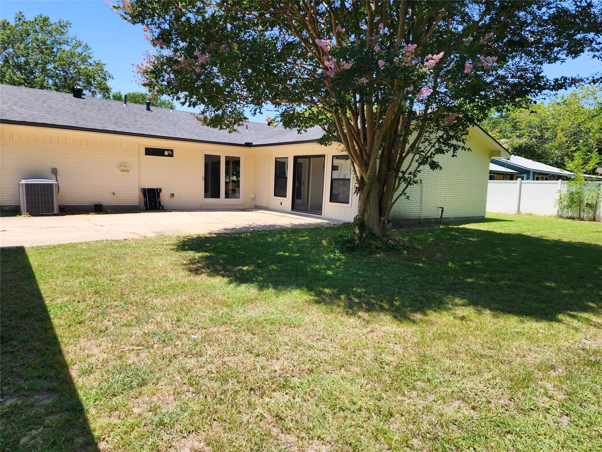 Expansive backyard featuring a concrete patio, mature tree, manicured lawn, and white privacy fencing