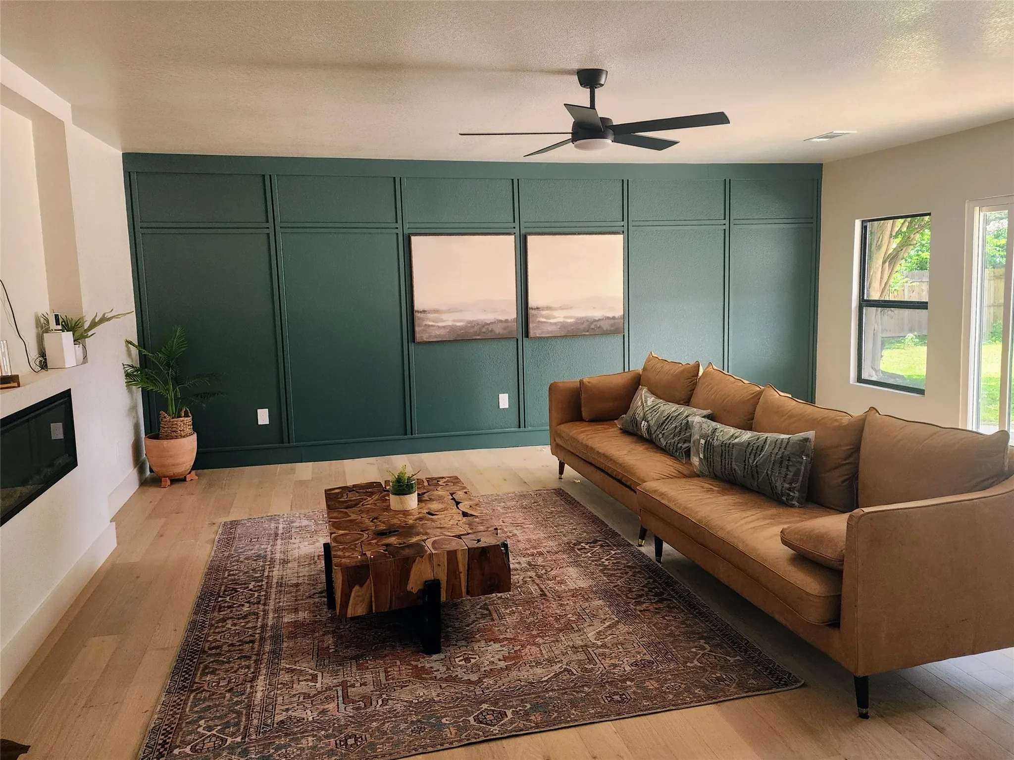 Accent wall with panel molding, light wood-finish flooring, recessed electric fireplace, black ceiling fan with integrated light, and large windows