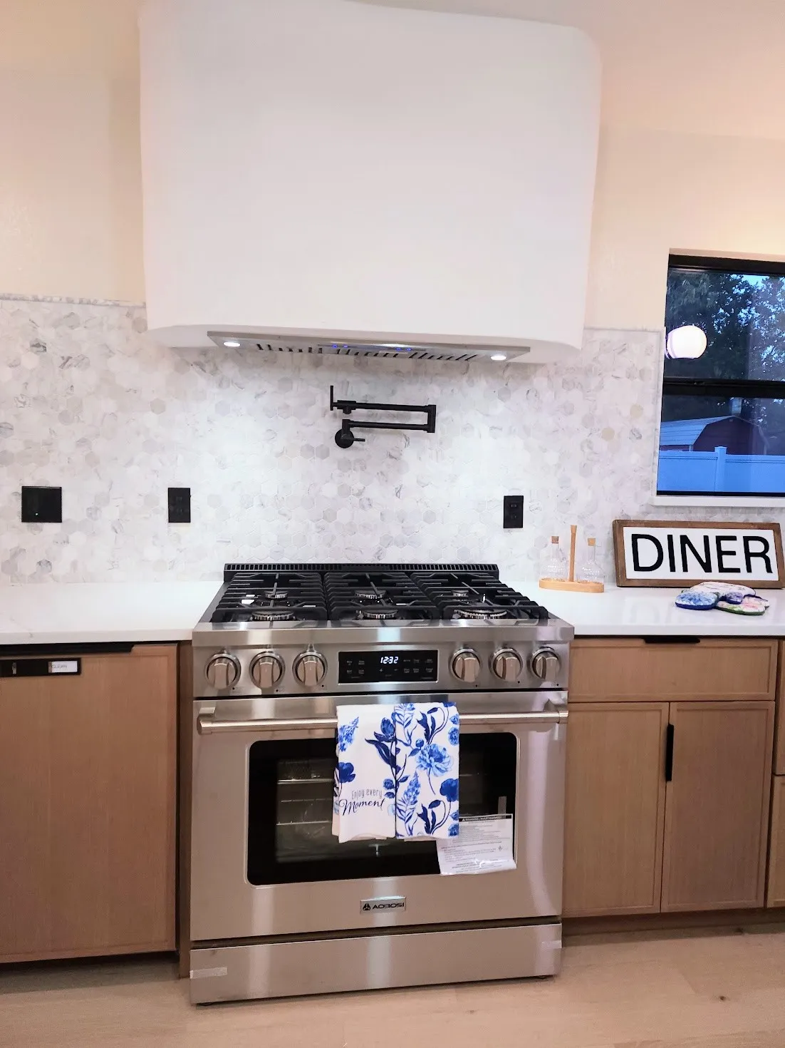 Stainless steel range with a pot filler, white curved range hood, tiled backsplash, light wood-finish cabinetry, and light countertops