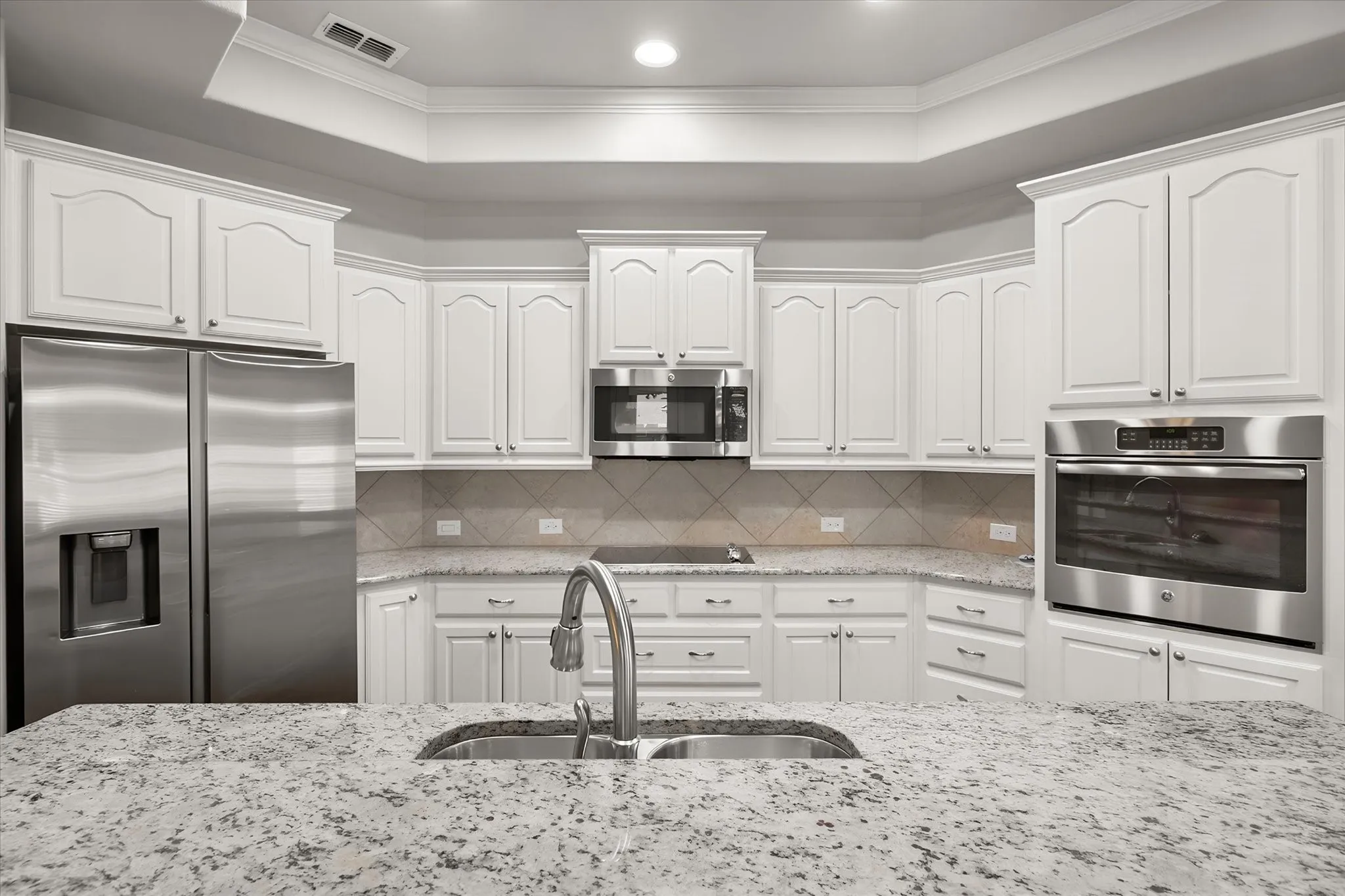 Gourmet kitchen showcases an abundance of custom white shaker cabinetry, granite countertops, stainless steel appliances and a coordinating tile backsplash. The spacious granite island features a double-basin sink and breakfast bar, creating an ideal workspace for cooking and entertaining. Accent lighting above the cabinets adds warmth and ambiance. The stainless refrigerator may convey with the home.