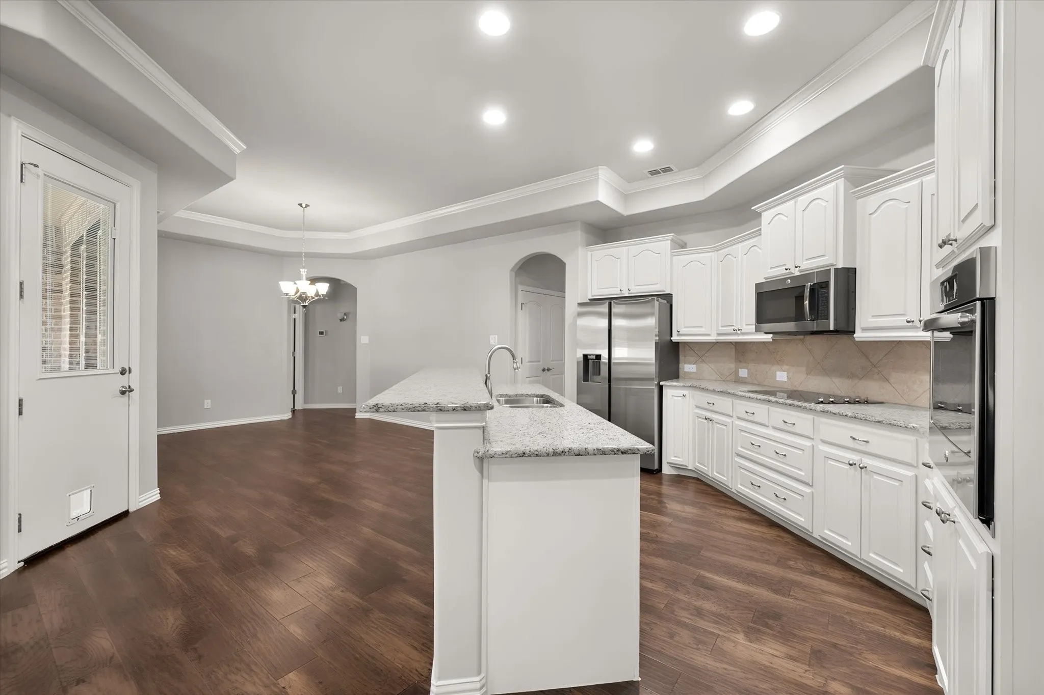 Spacious custom kitchen designed for both everyday living and entertaining, featuring an oversized granite breakfast bar with seating, abundant custom cabinetry, stainless steel appliances and a tile backsplash. The open-concept layout flows seamlessly into the dining and living areas, while rich wood flooring, crown molding, arched doorways and recessed lighting enhance the home’s upscale garden-home design.