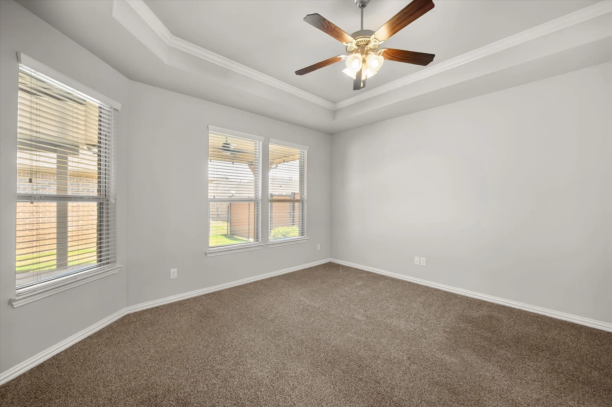 Primary bedroom with plush carpeting, a tray ceiling with crown molding, ceiling fan, and multiple windows providing abundant natural light.