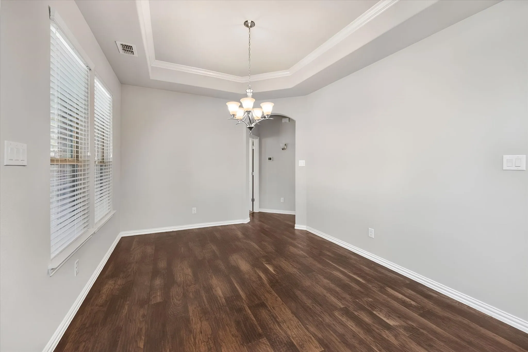 Elegant dining room designed for entertaining, featuring rich wood flooring, an architectural inset tray ceiling with crown molding, and abundant natural light through expansive windows with two inch blinds. An arched architectural opening connects the dining space to the main living areas, while the convenient patio door creates an effortless connection to outdoor living and entertaining.