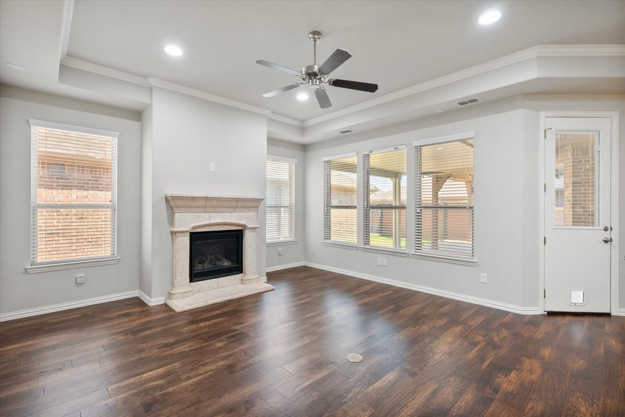 Spacious living room in this luxury custom garden home showcases rich wood flooring, soaring ceilings and walls of windows that fill the space with abundant natural light. A beautiful gas-log fireplace with a substantial stone surround creates an inviting focal point, while recessed lighting, crown molding and a modern ceiling fan add comfort and architectural appeal.
