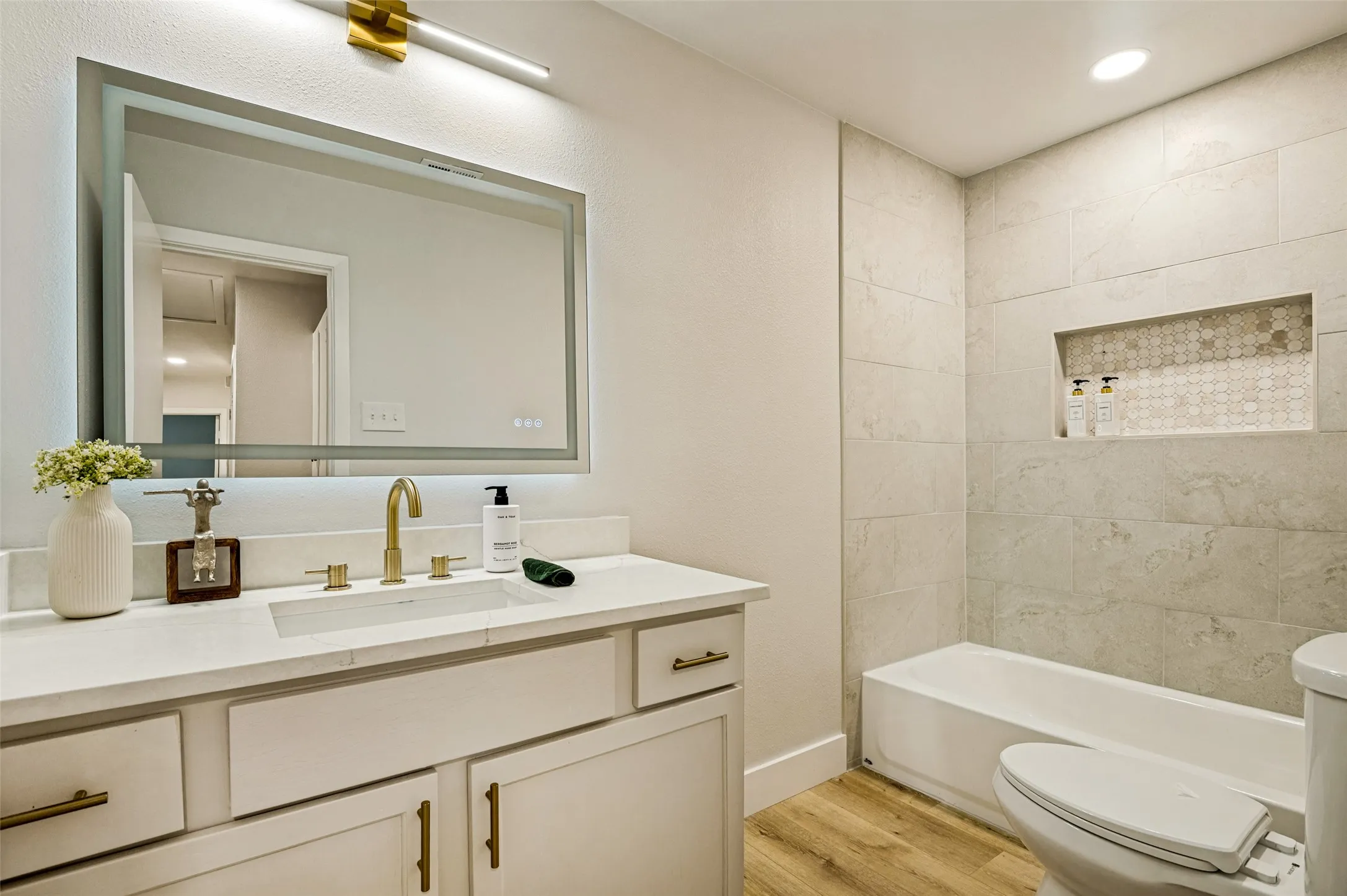 Bathroom featuring a rectangular undermount sink, a large illuminated mirror, a built-in tub, and a tiled shower niche with decorative inlay
