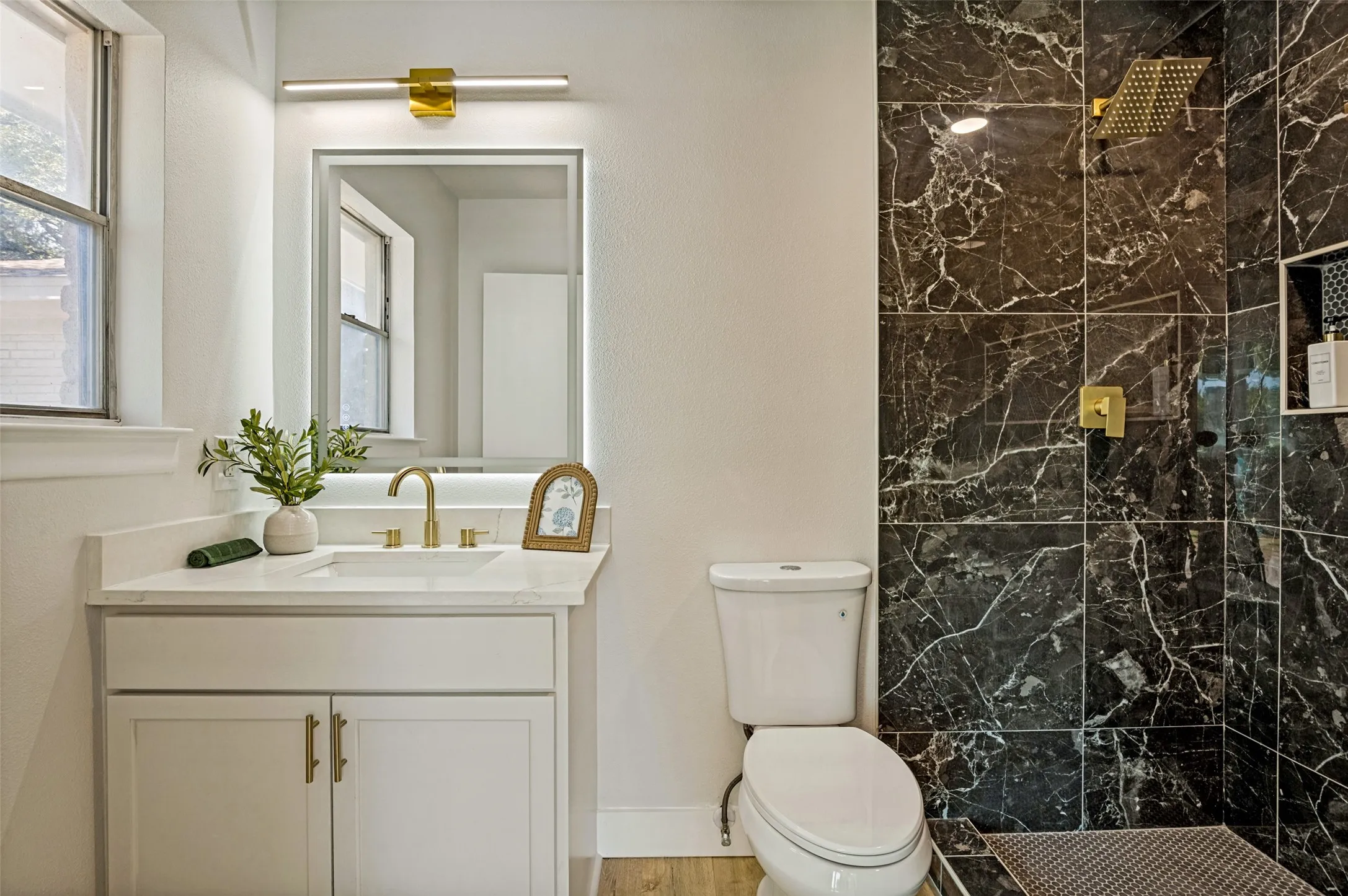 Black marble-style shower tile, gold-tone shower fixtures, white vanity with gold-tone hardware, white countertop with integrated sink, and gold-tone faucet
