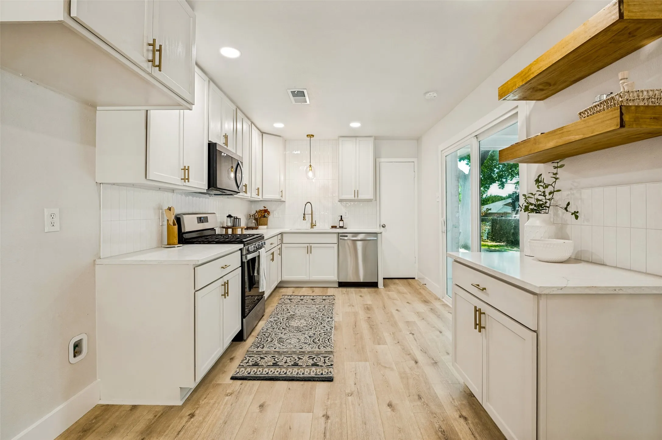 White shaker cabinetry with gold-tone hardware, white countertops, subway tile backsplash, stainless steel appliances, and light wood-finish flooring