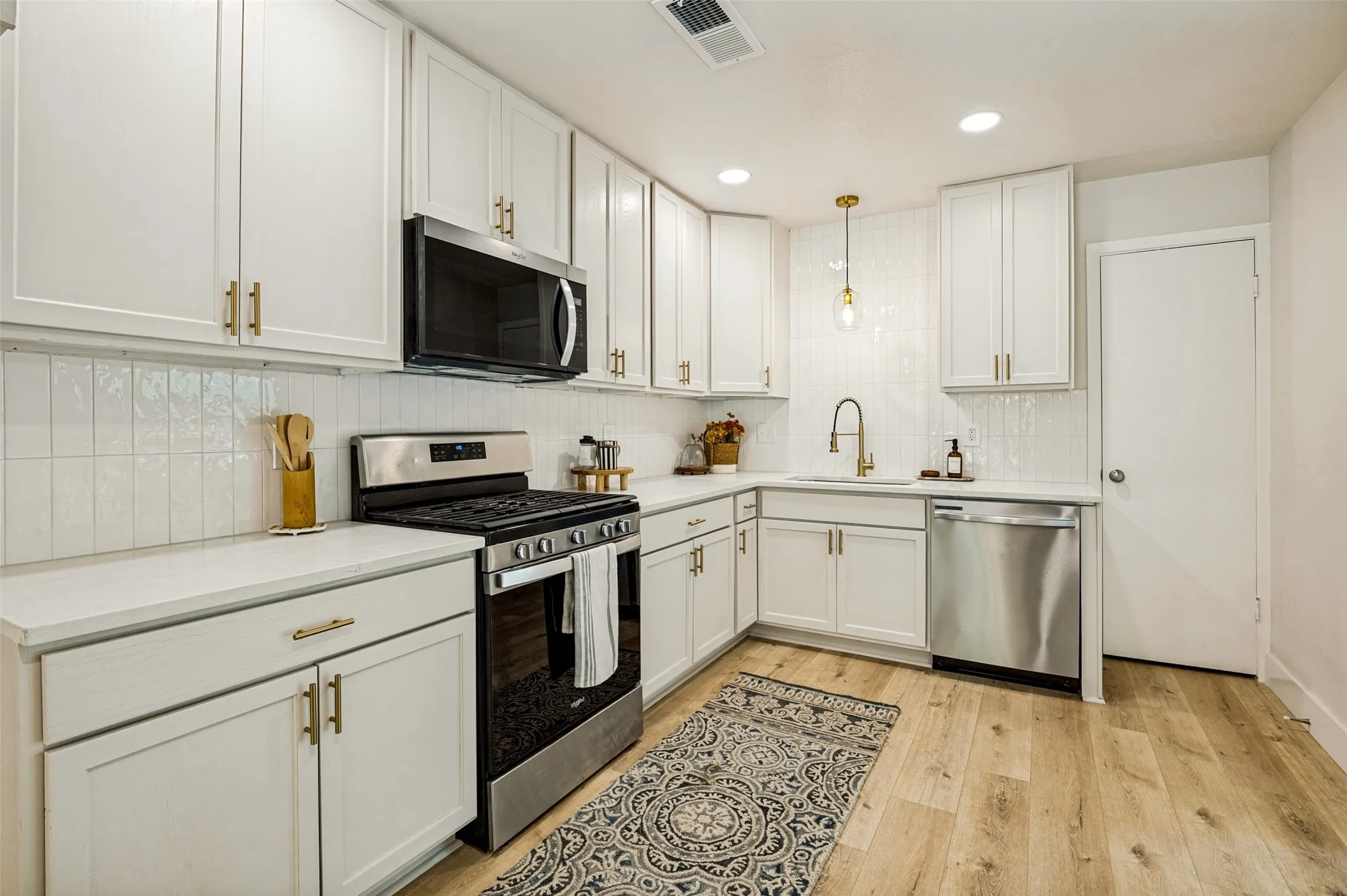 White cabinetry with gold-tone hardware, stainless steel appliances, white subway tile backsplash, light wood-finish flooring, and recessed lighting