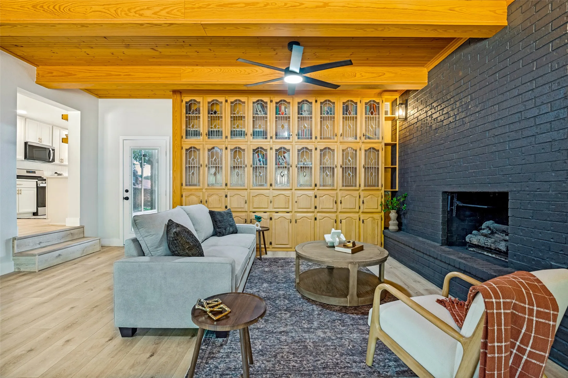 Exposed wood beams, built-in cabinetry with glass-front doors, a dark brick fireplace, a ceiling fan, and wood-finish flooring