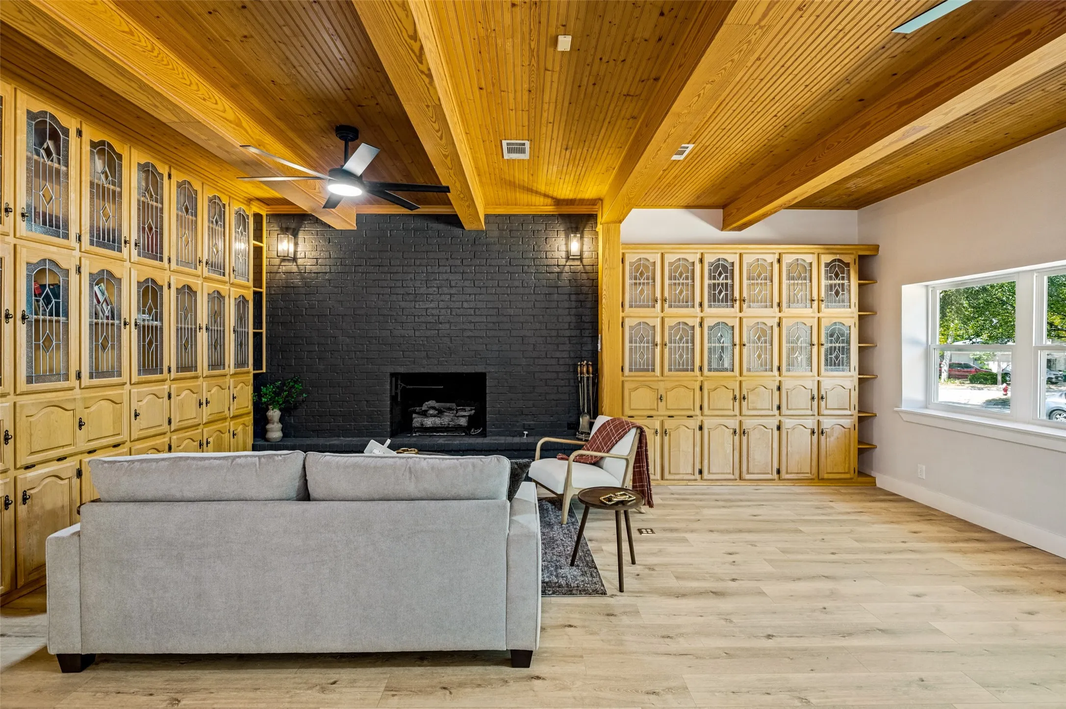 Wood-finish flooring, exposed wood beam ceiling, built-in cabinetry with glass inserts, black brick fireplace, and multiple windows