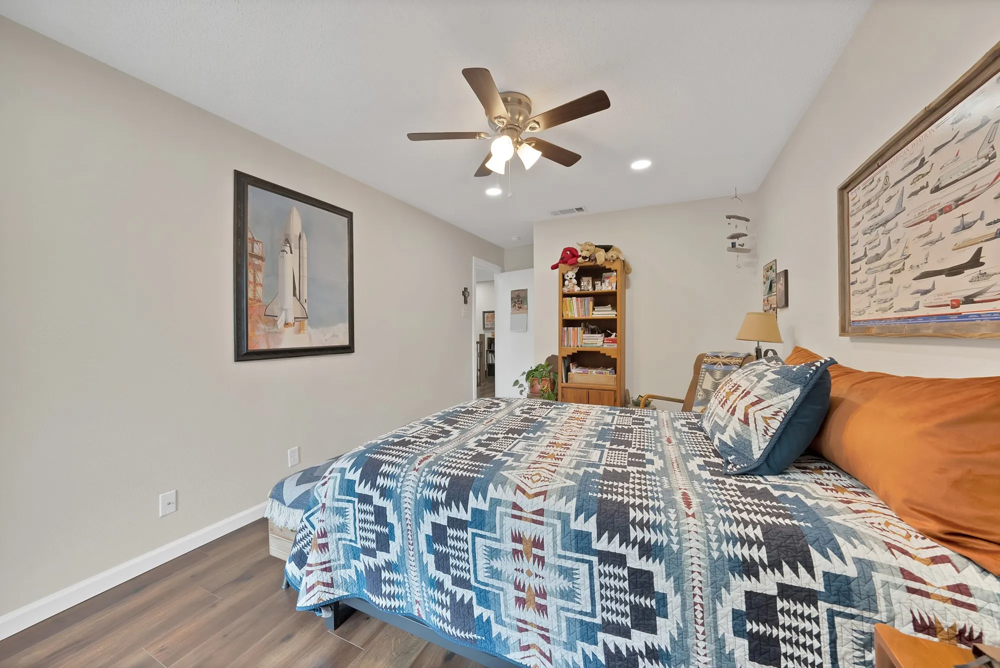 Bedroom — Wood look flooring with ceiling fan and bookshelf, adjoining hallway access.