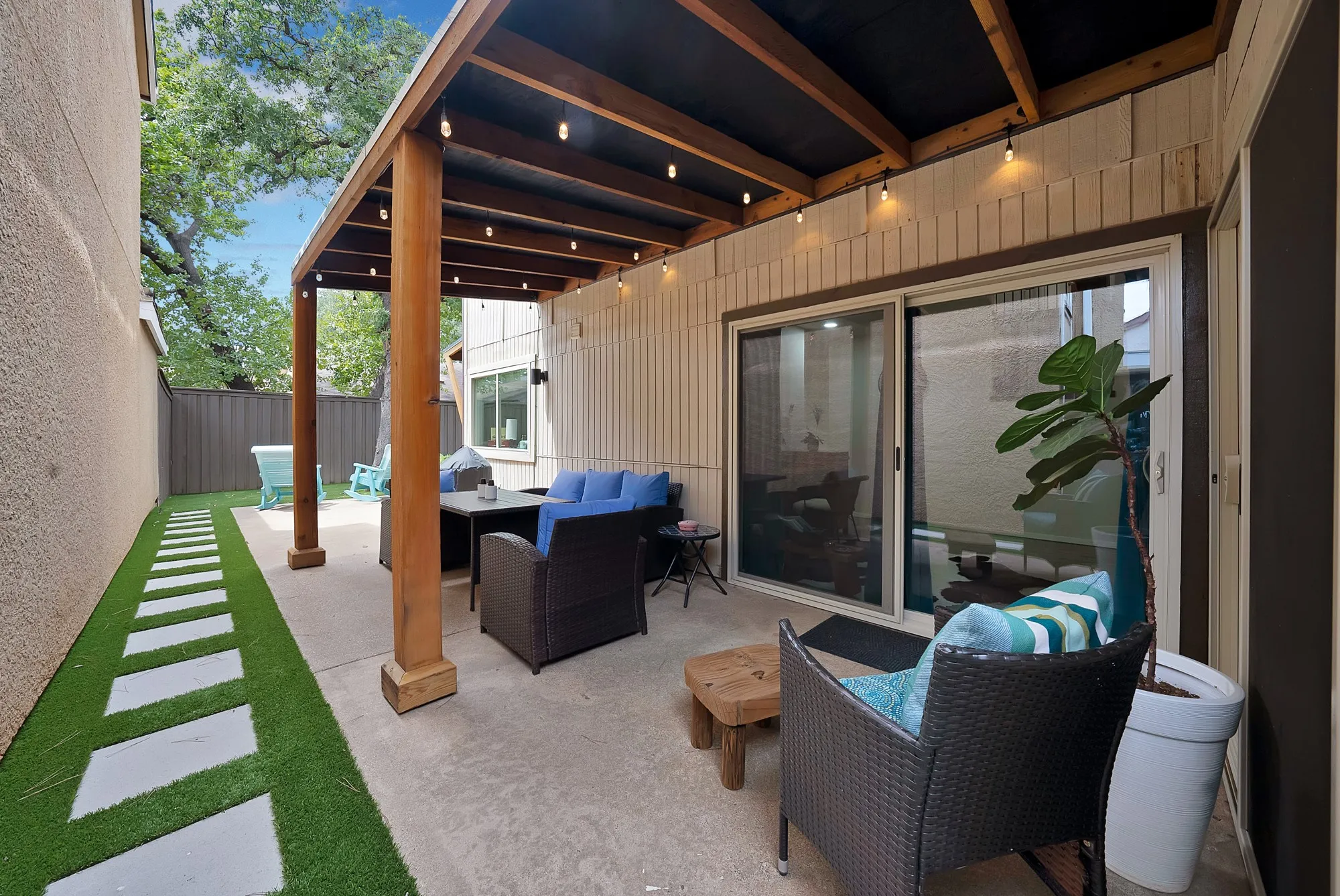 Covered patio — String lighted wood pergola over patio with wicker seating, sliding glass doors to interior, and turf pathway with pavers.