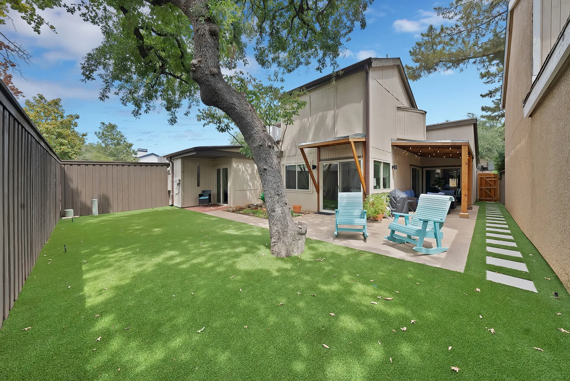 Rear exterior — Two story home with covered patio, turf lawn, mature tree, and teal Adirondack rocking chairs.