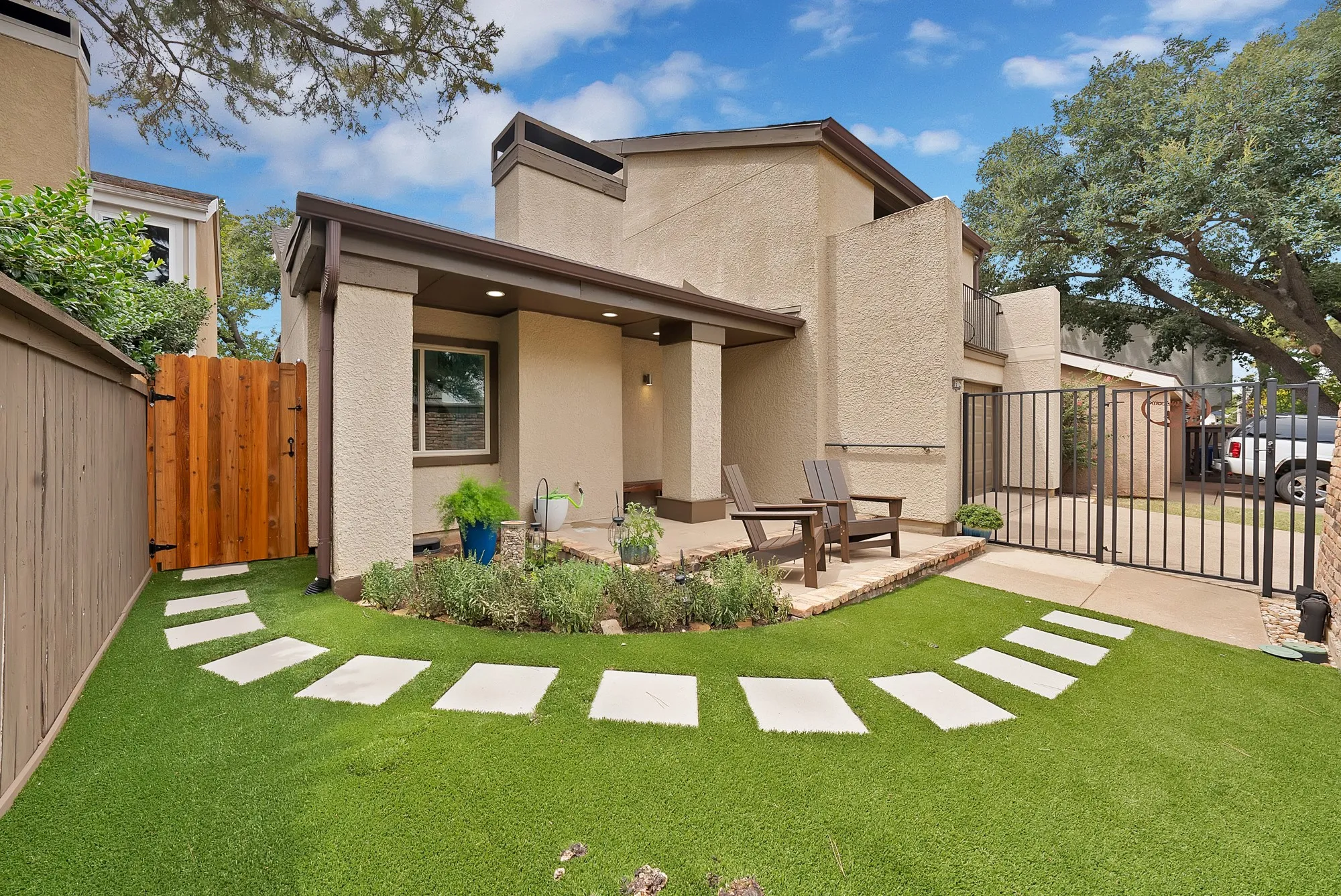 Two story stucco home with covered patio, Adirondack chairs, wrought iron gate, and turf lawn with stepping stone path.