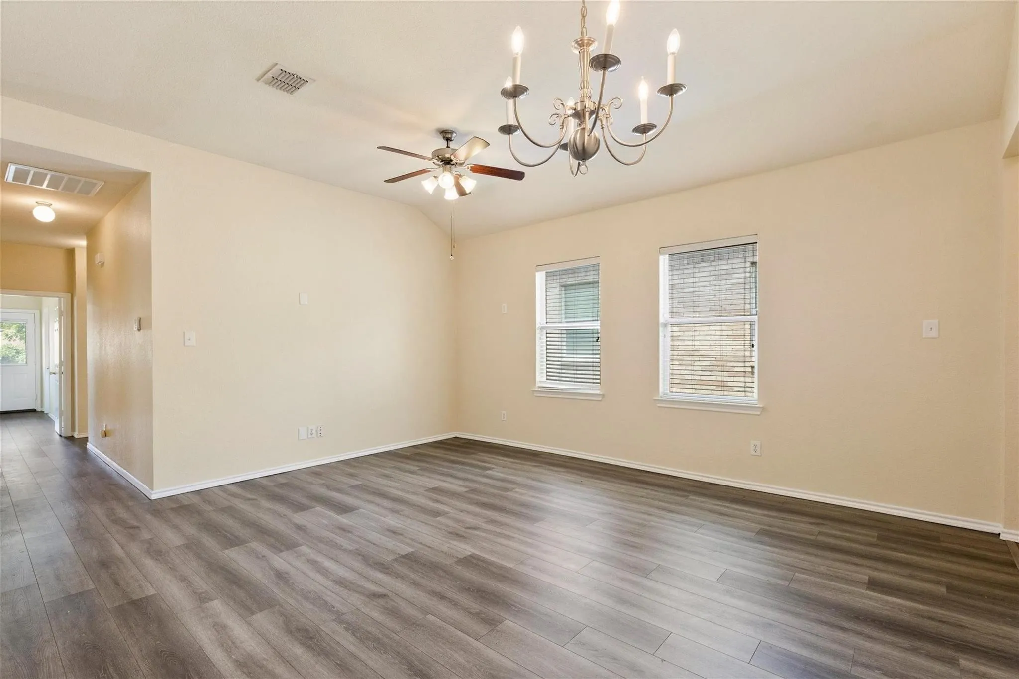 Spacious room featuring wood-finish flooring, two large windows with blinds, a ceiling fan, and a decorative chandelier