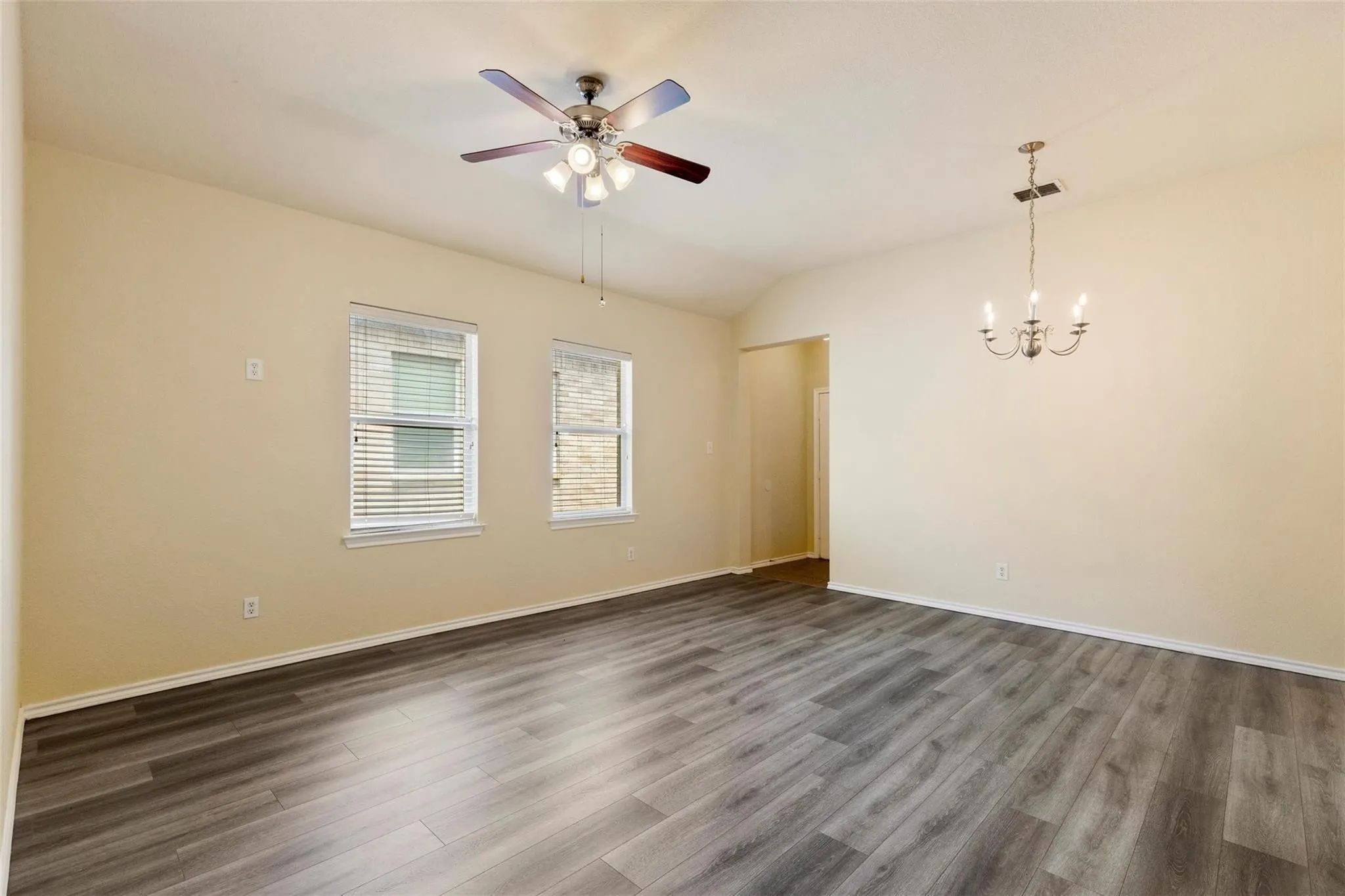 Vaulted ceiling, wood-finish flooring, two windows with blinds, ceiling fan with integrated lighting, and a chandelier