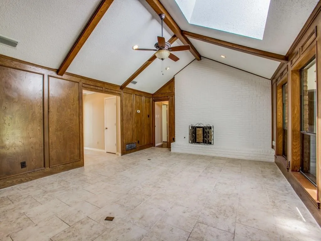 Vaulted ceiling with exposed wood beams, skylight, white-painted brick fireplace, wood paneling, and large format tile flooring