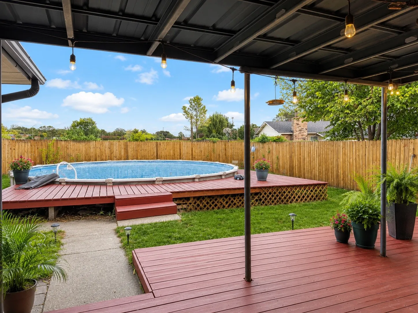 Vertually stagedCovered patio with string lighting, red-stained wood decking, and an above-ground pool with surrounding wood deck