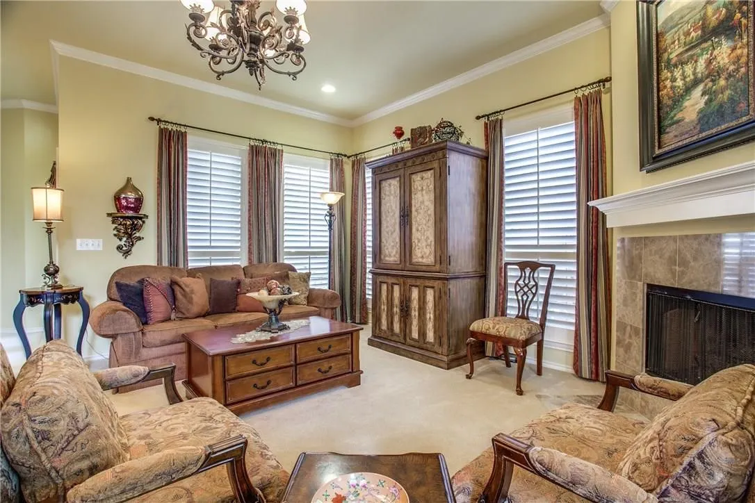 Carpeted living area featuring a stone-surround fireplace with a white mantel, multiple windows with plantation shutters, an ornate chandelier, and crown molding