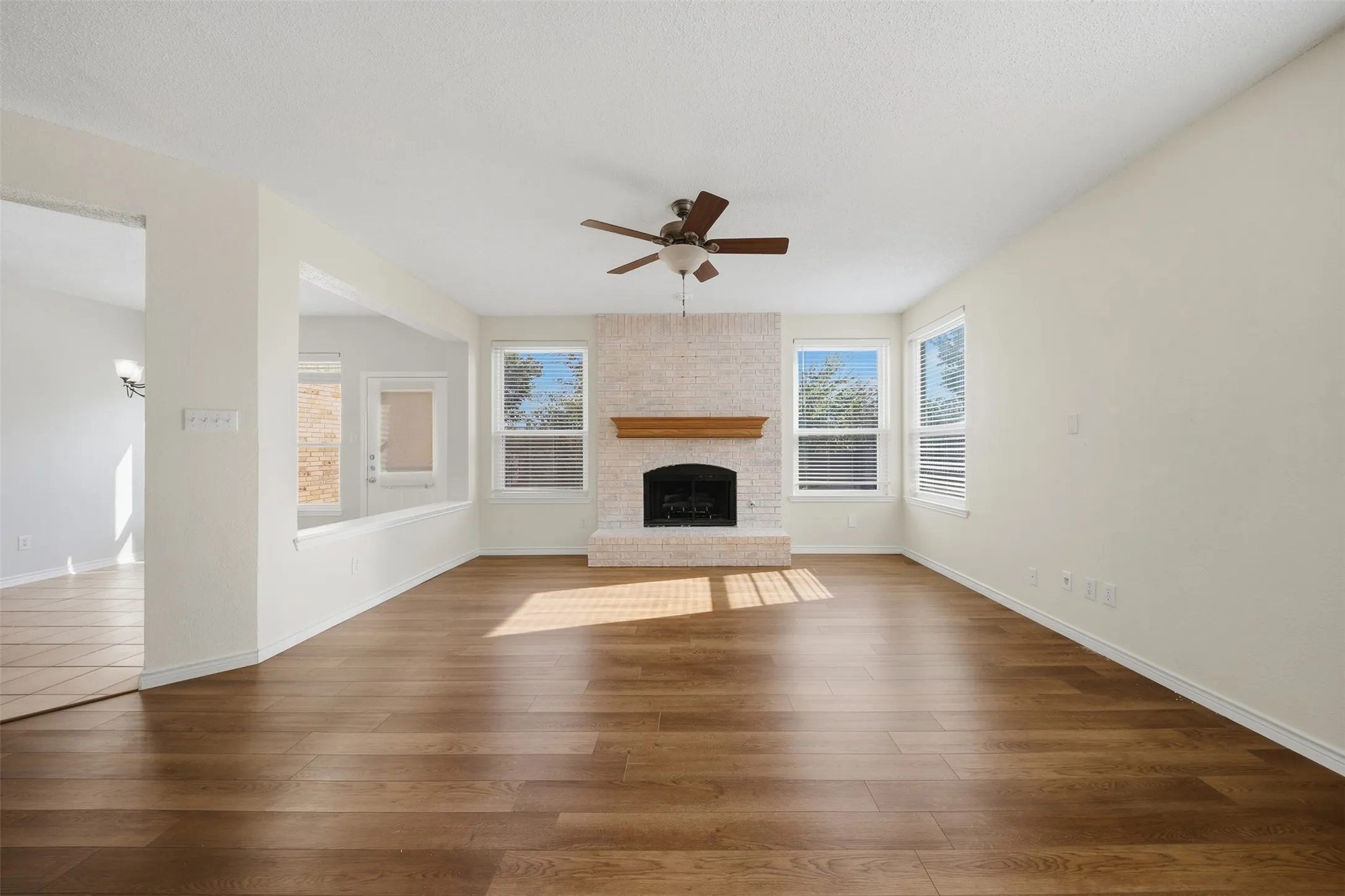 Wood-finish flooring, white brick fireplace with wood mantel, ceiling fan, multiple windows with blinds, and recessed half-wall opening