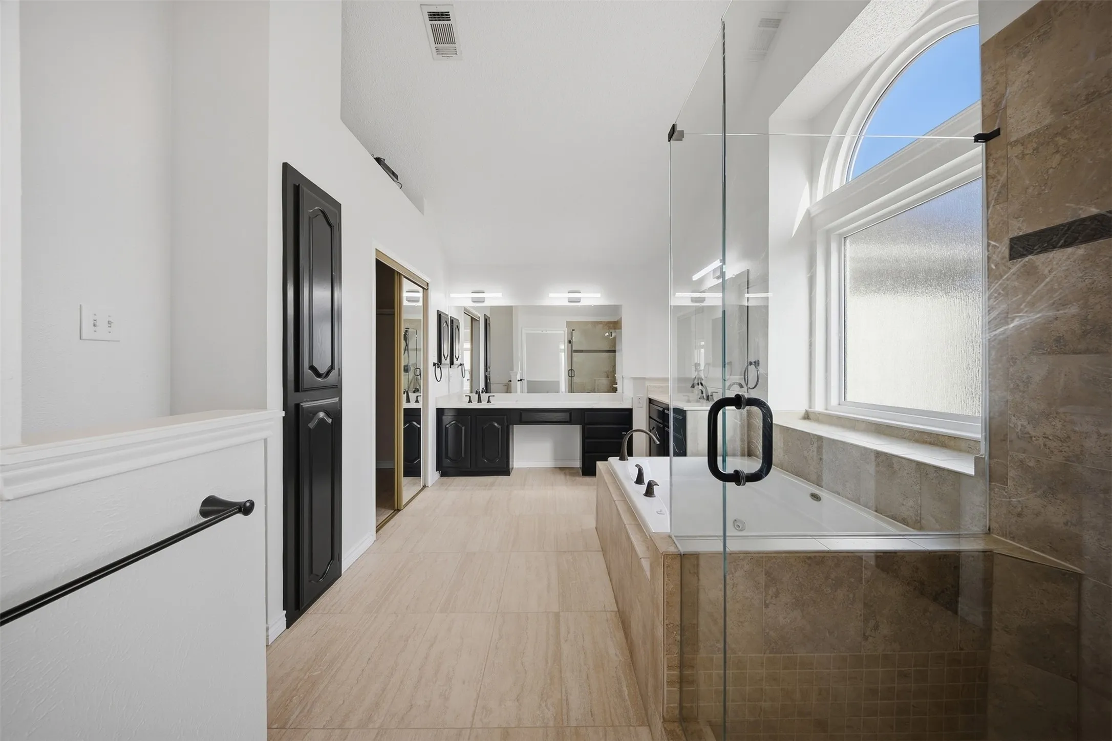 Expansive bathroom featuring light-toned tile flooring, a built-in jetted tub with tile surround, a glass-enclosed shower, dual vanity with dark cabinetry, and a large arched window