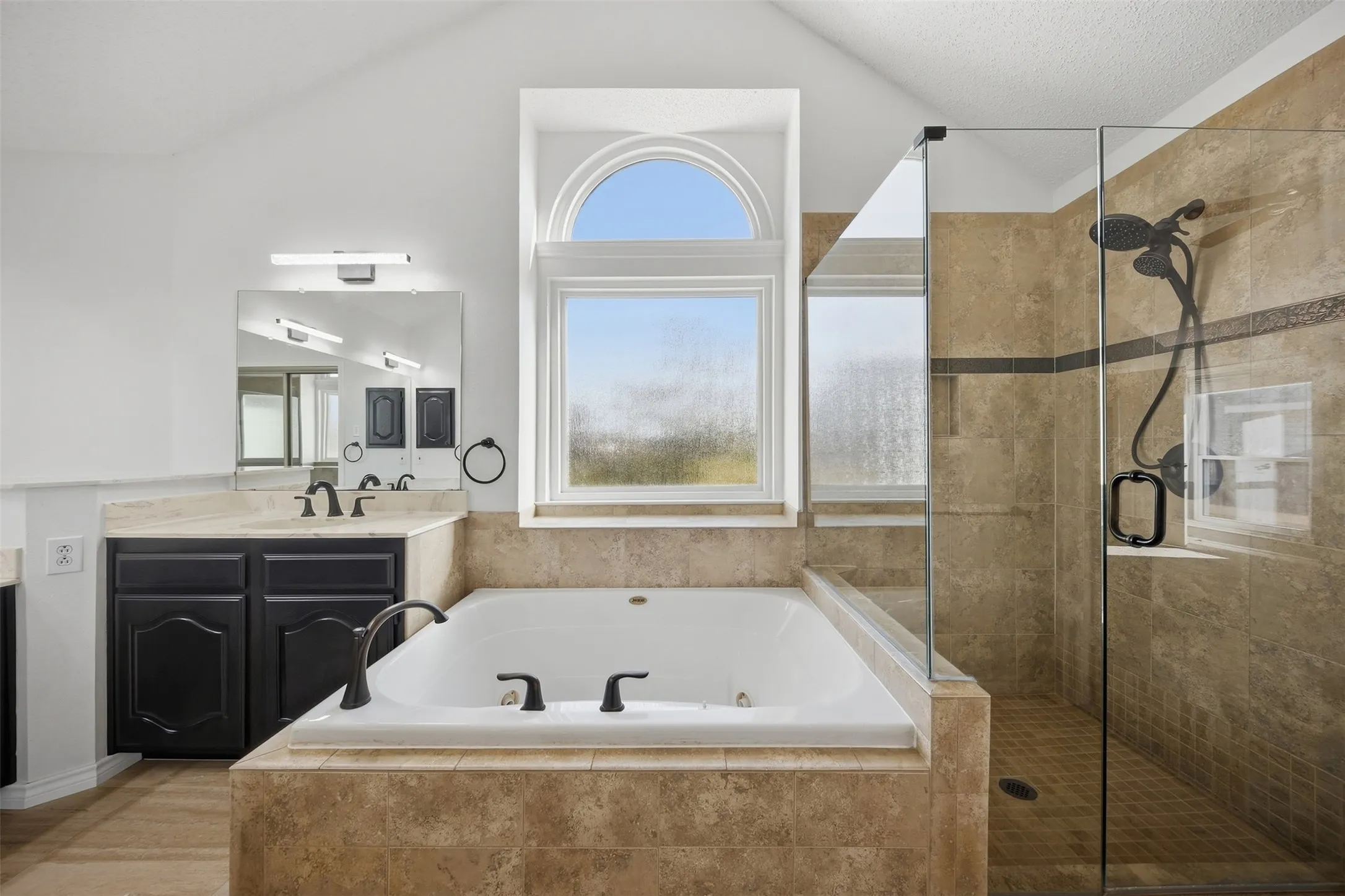 Tile-surrounded soaking tub, separate glass-enclosed shower, dual-sink vanity with dark cabinetry, and an arched transom window