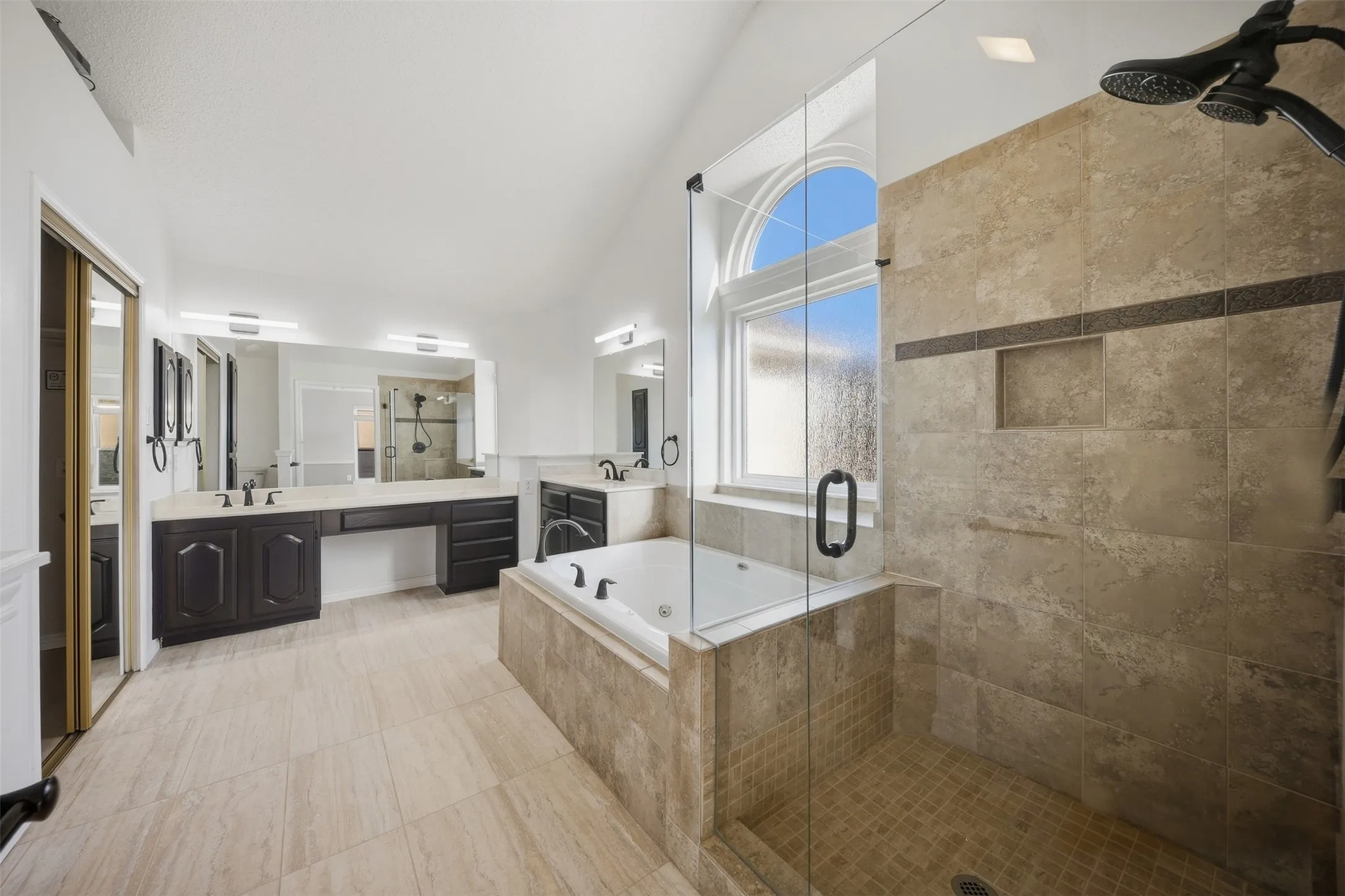 Expansive bathroom featuring dual vanities, a deep soaking tub, a separate glass-enclosed shower, a large arched window, and tile flooring