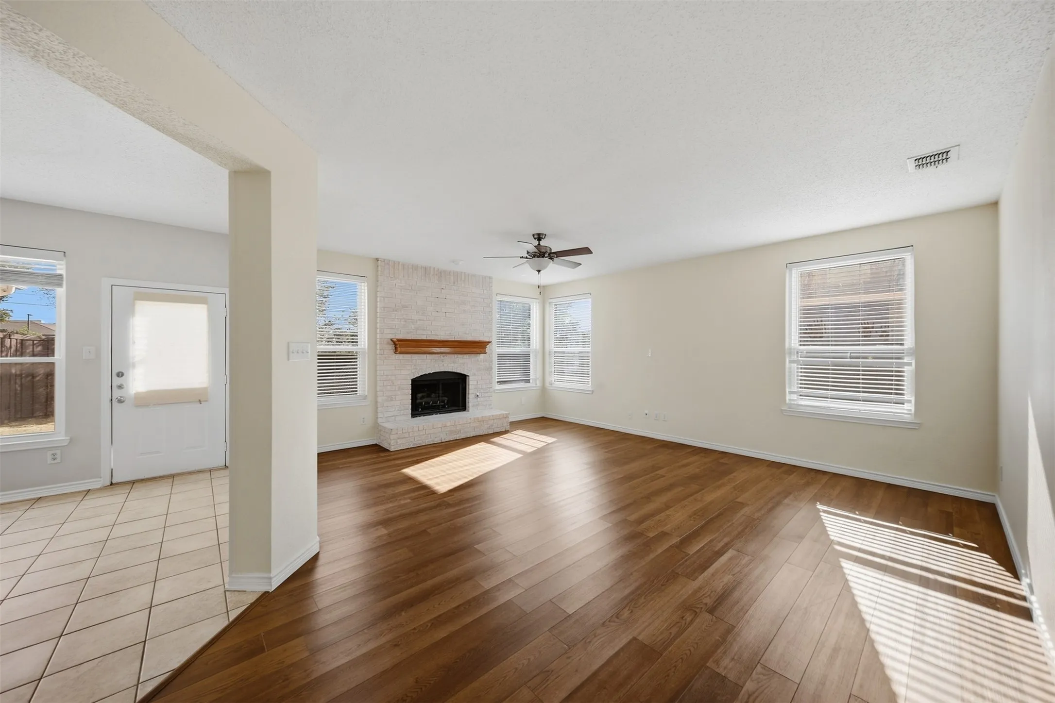 Spacious living area featuring wood-finish flooring, a white brick fireplace with a wood mantle, and a ceiling fan