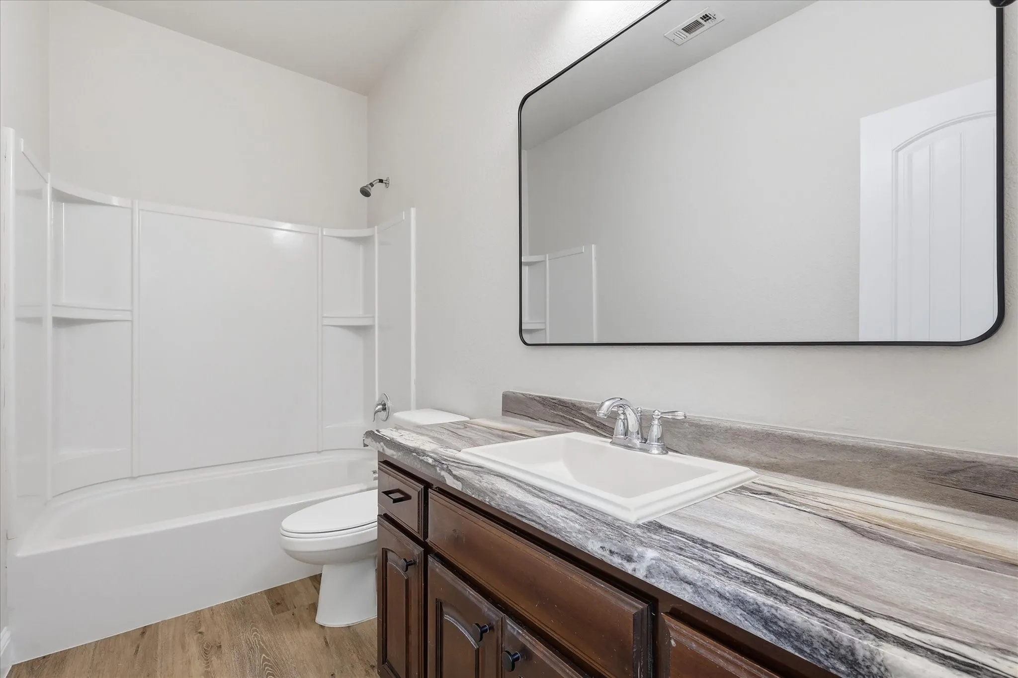 Wood-finish flooring, dark wood vanity with white rectangular sink, framed rectangular mirror, and a white tub with shower