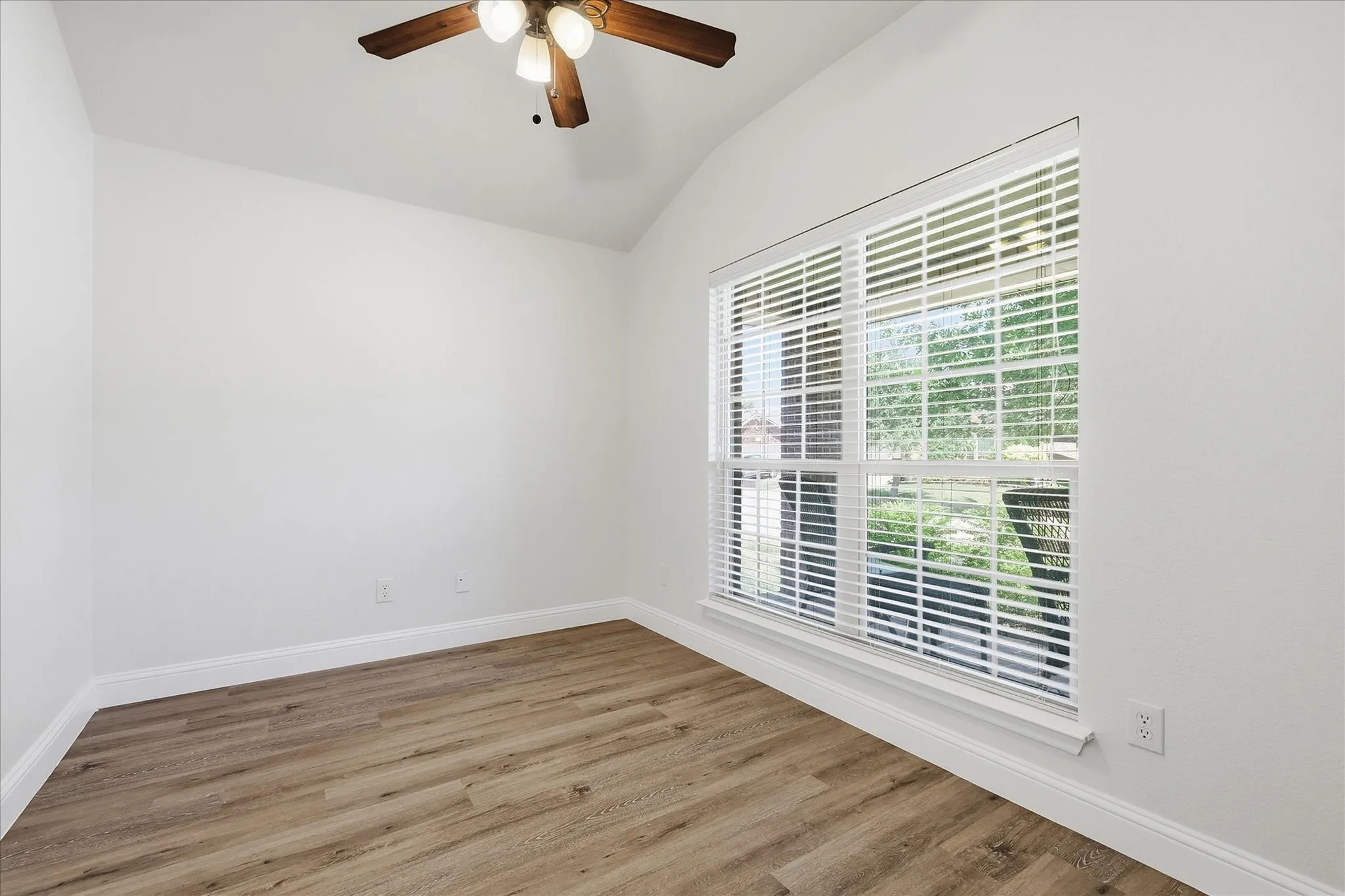 Vaulted ceiling, ceiling fan with light fixture, wood-finish flooring, white walls, and a window with blinds