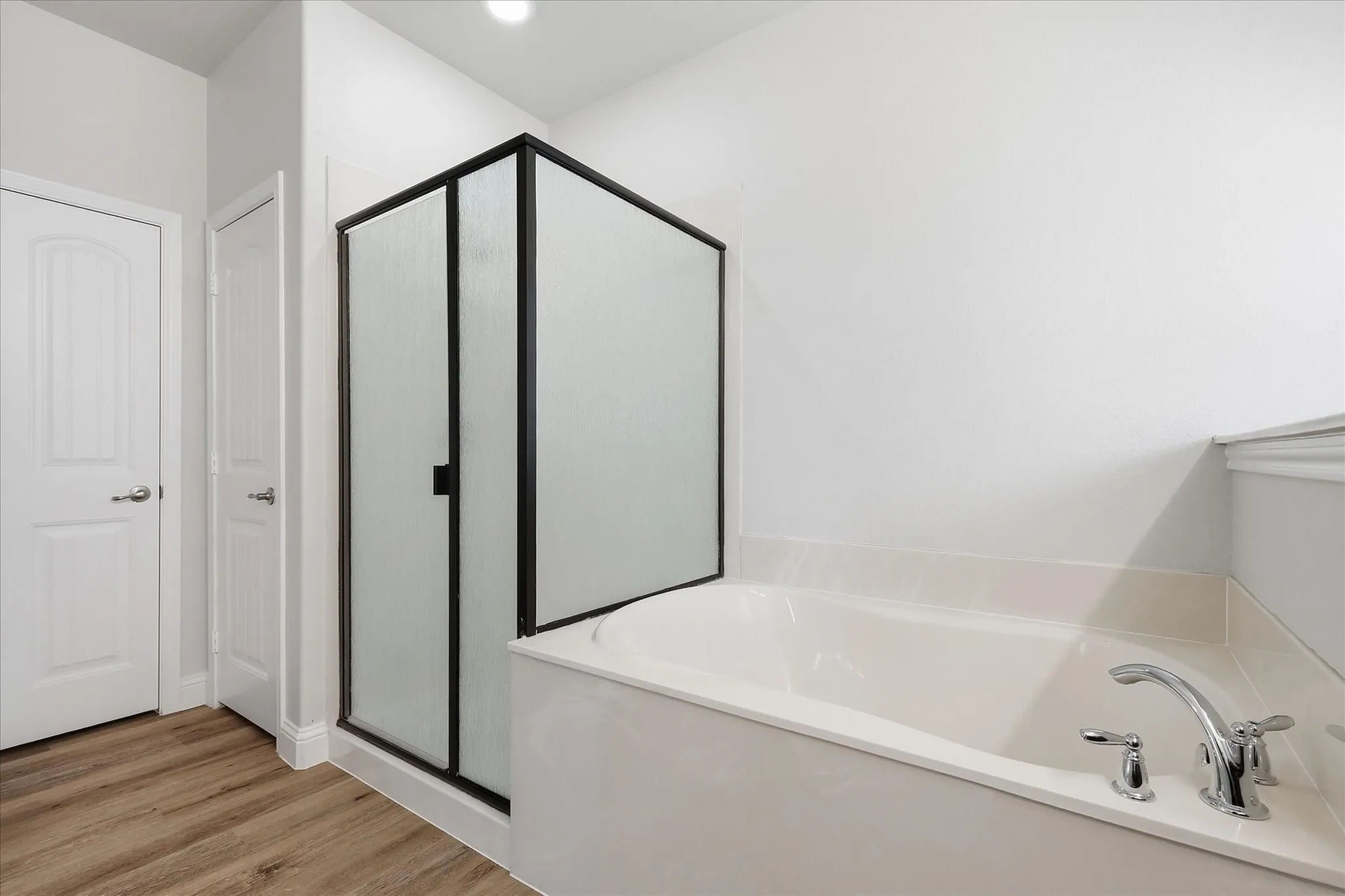 Soaking tub with chrome fixtures, enclosed shower with a black frame and frosted glass, wood-finish flooring, and white paneled doors