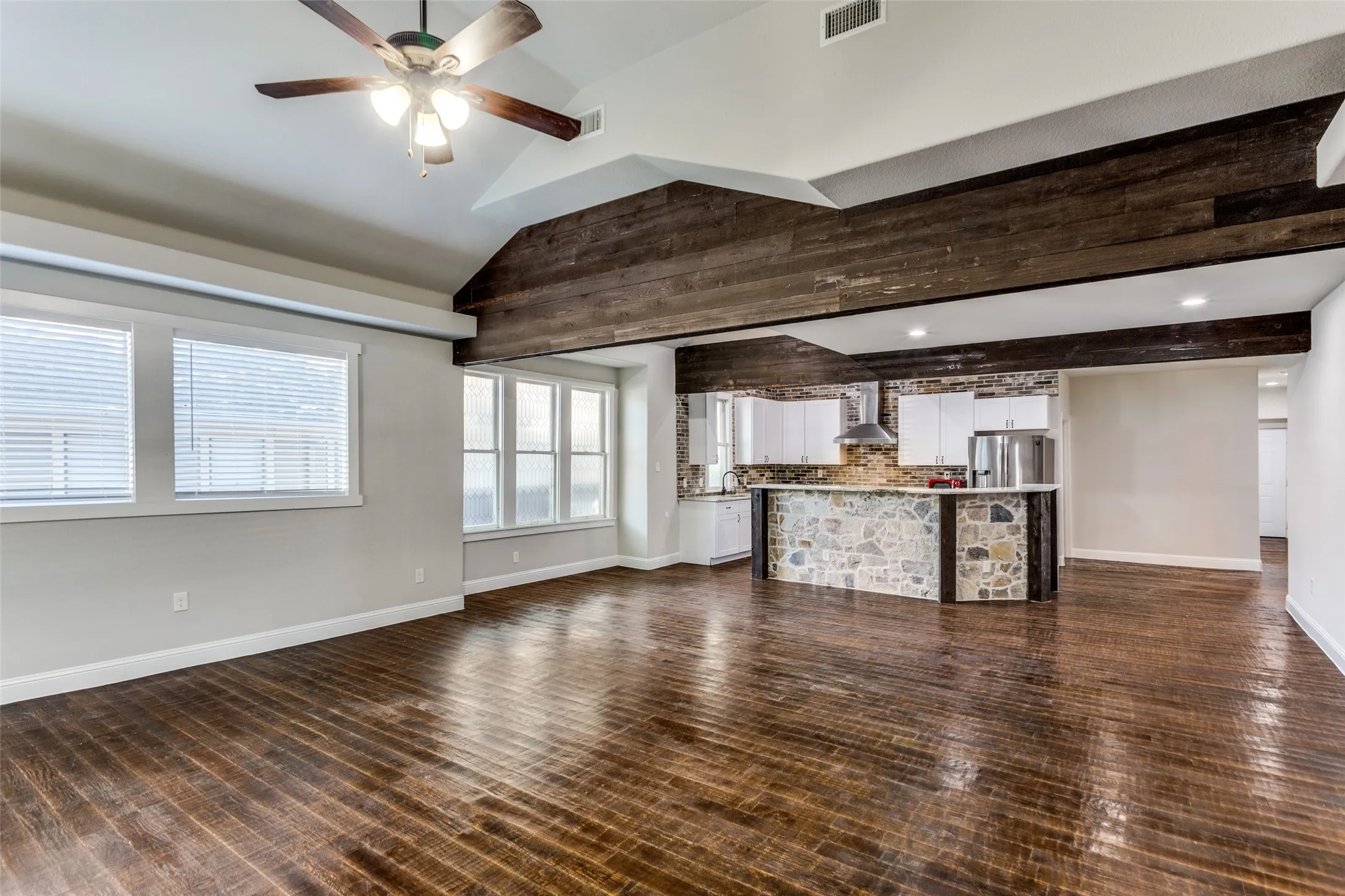 Vaulted ceiling with wood-finish beams, dark wood-finish flooring, multiple windows with blinds, kitchen island with stone detailing, and stainless steel range hood