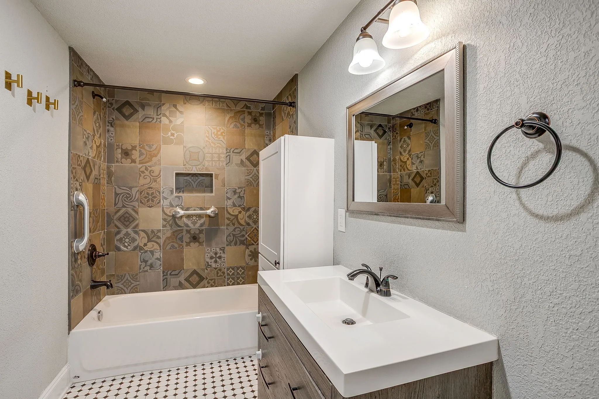 Patterned tile shower surround with integrated niche, white bathtub with matte black fixtures, wood-finish vanity with an integrated sink, and black and white mosaic floor tile