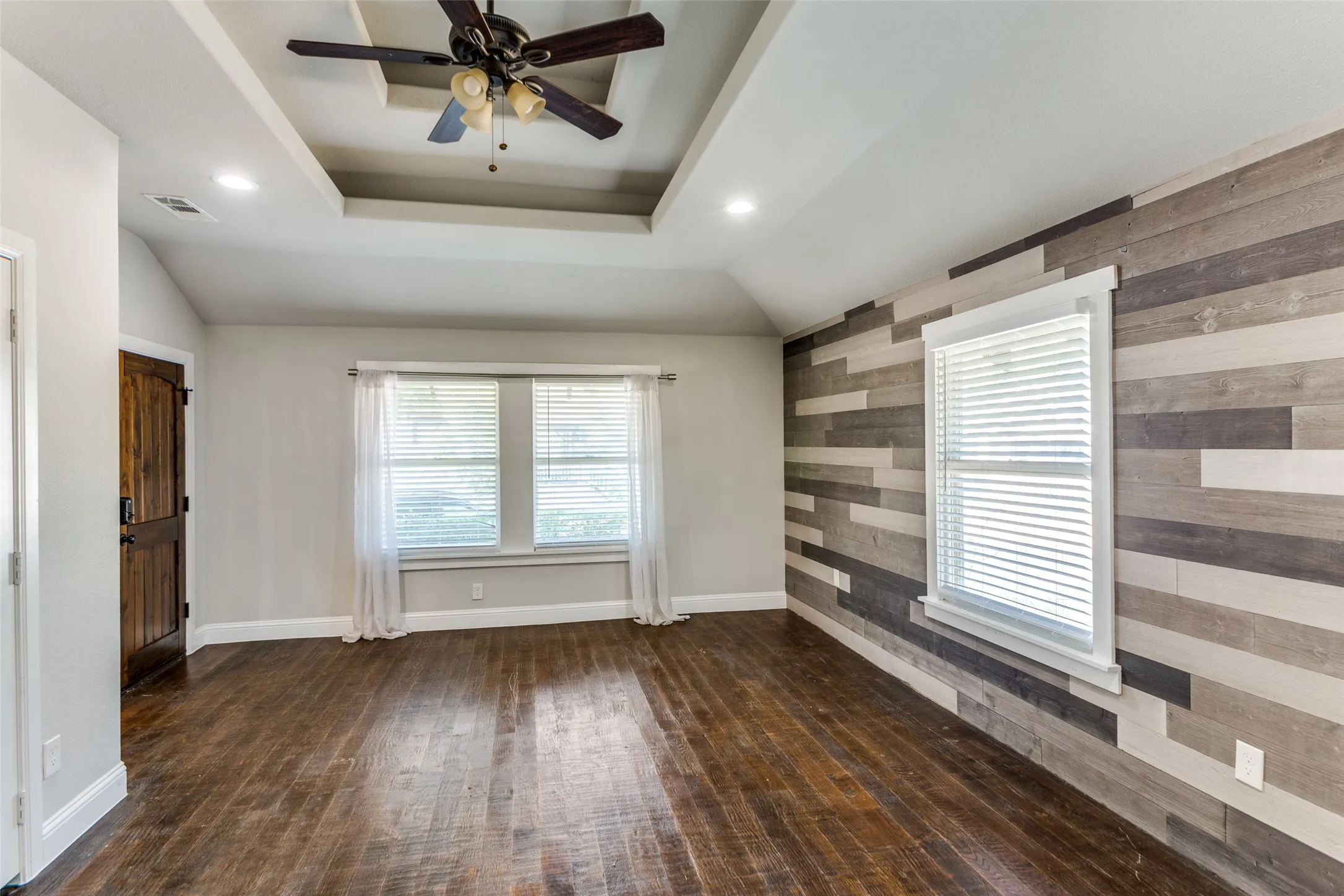 Tray ceiling with recessed lighting and ceiling fan, wood-finish flooring, wood plank accent wall, and double windows with blinds